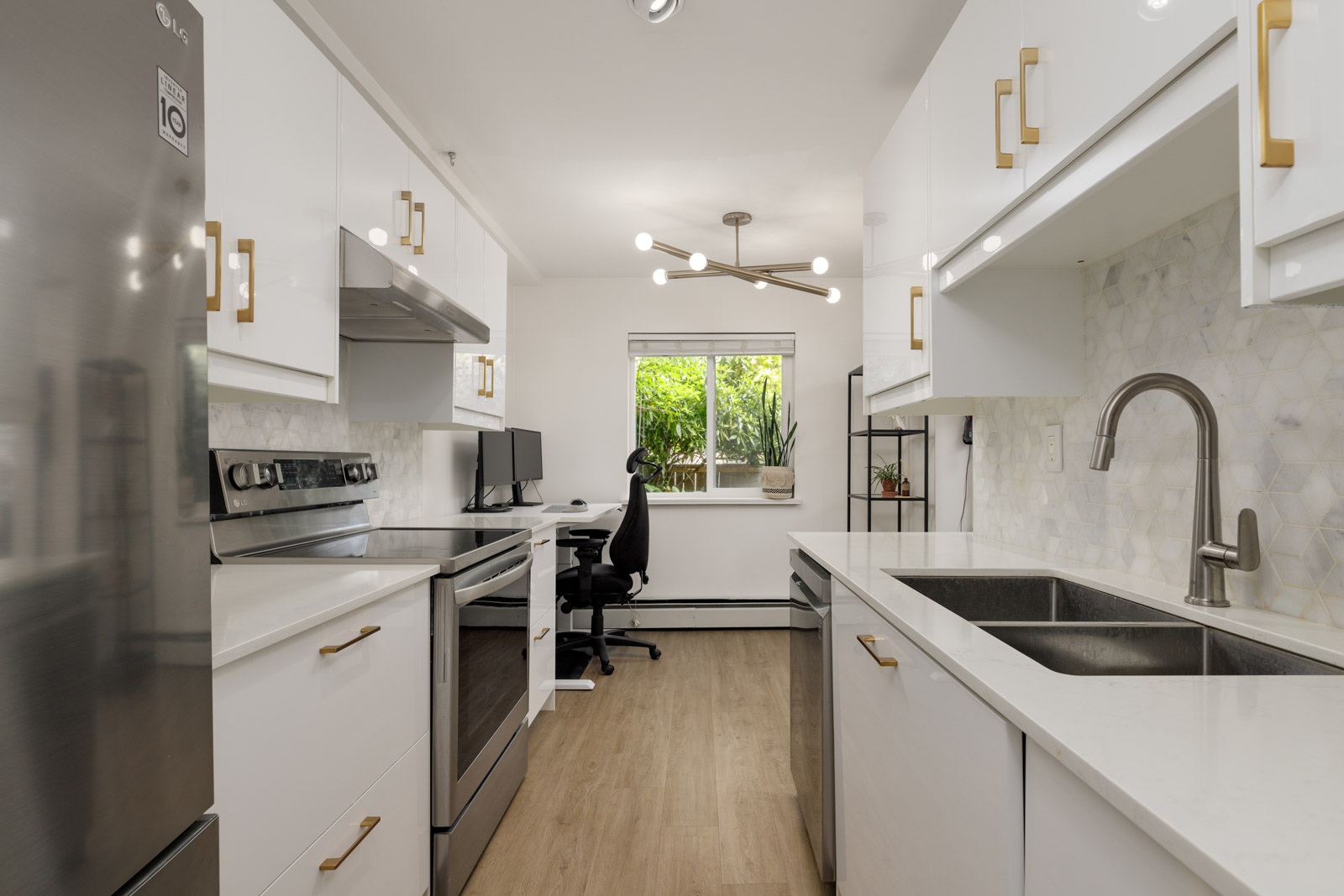Modern white kitchen with stainless steel appliances, gold handles, double sink, wood floor, and a desk with office chair by a window with plants.