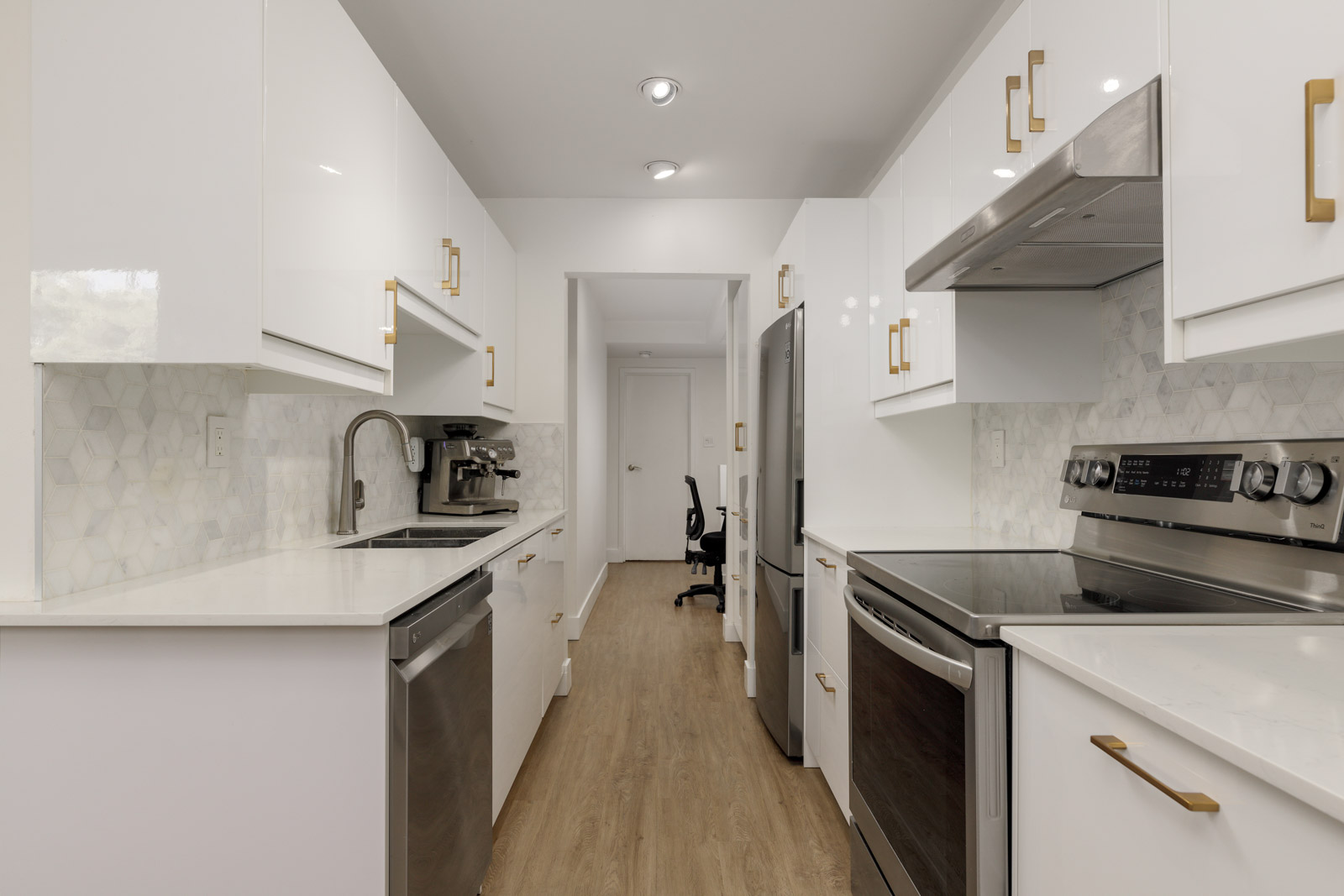 Modern galley kitchen with white cabinets, stainless steel appliances, gold hardware, light wood flooring, and a hexagon tile backsplash. An office chair is visible in the distant room.