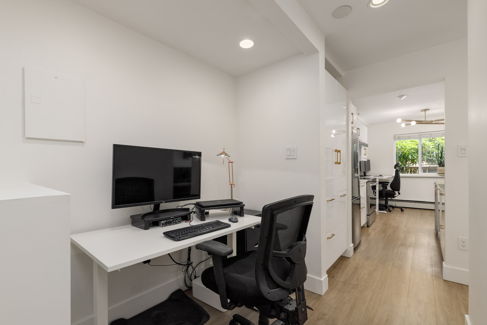 A home office setup with a computer, keyboard, mouse, and desk chair in a white room, adjacent to a hallway leading to a kitchen area with wooden flooring and large windows.
