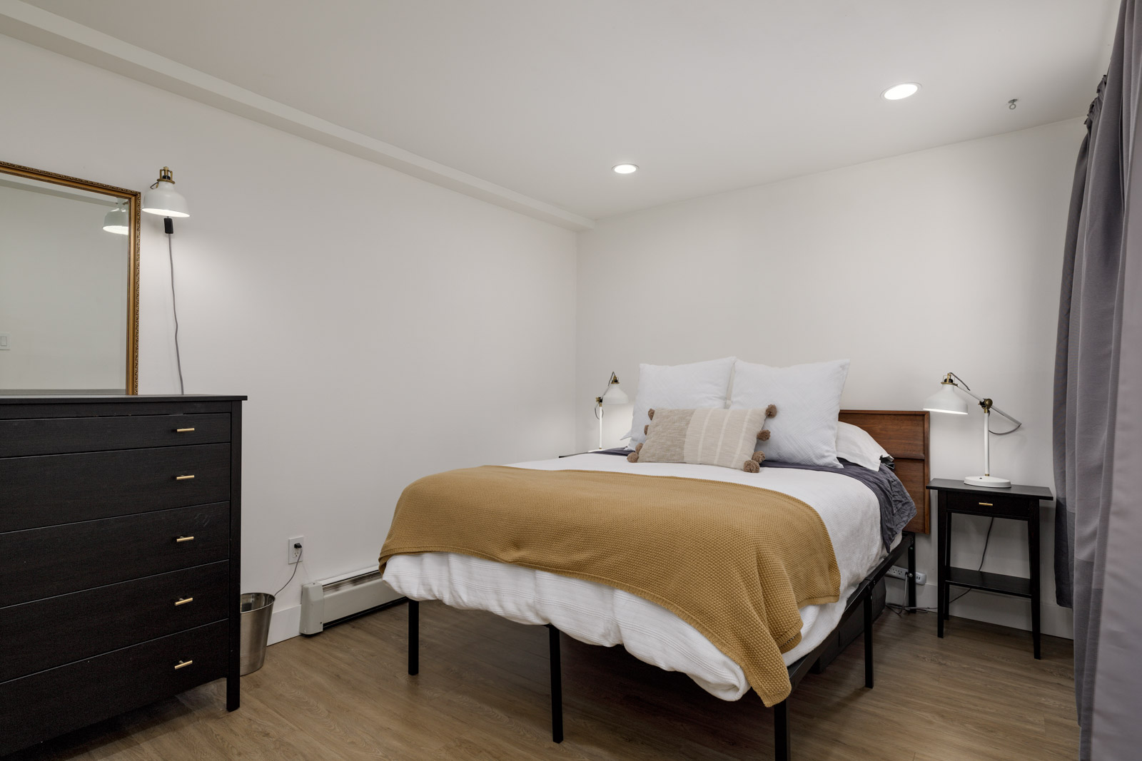 A neatly made bed with white and beige bedding sits in a simple bedroom with a black dresser, a small black desk, and neutral walls and flooring.