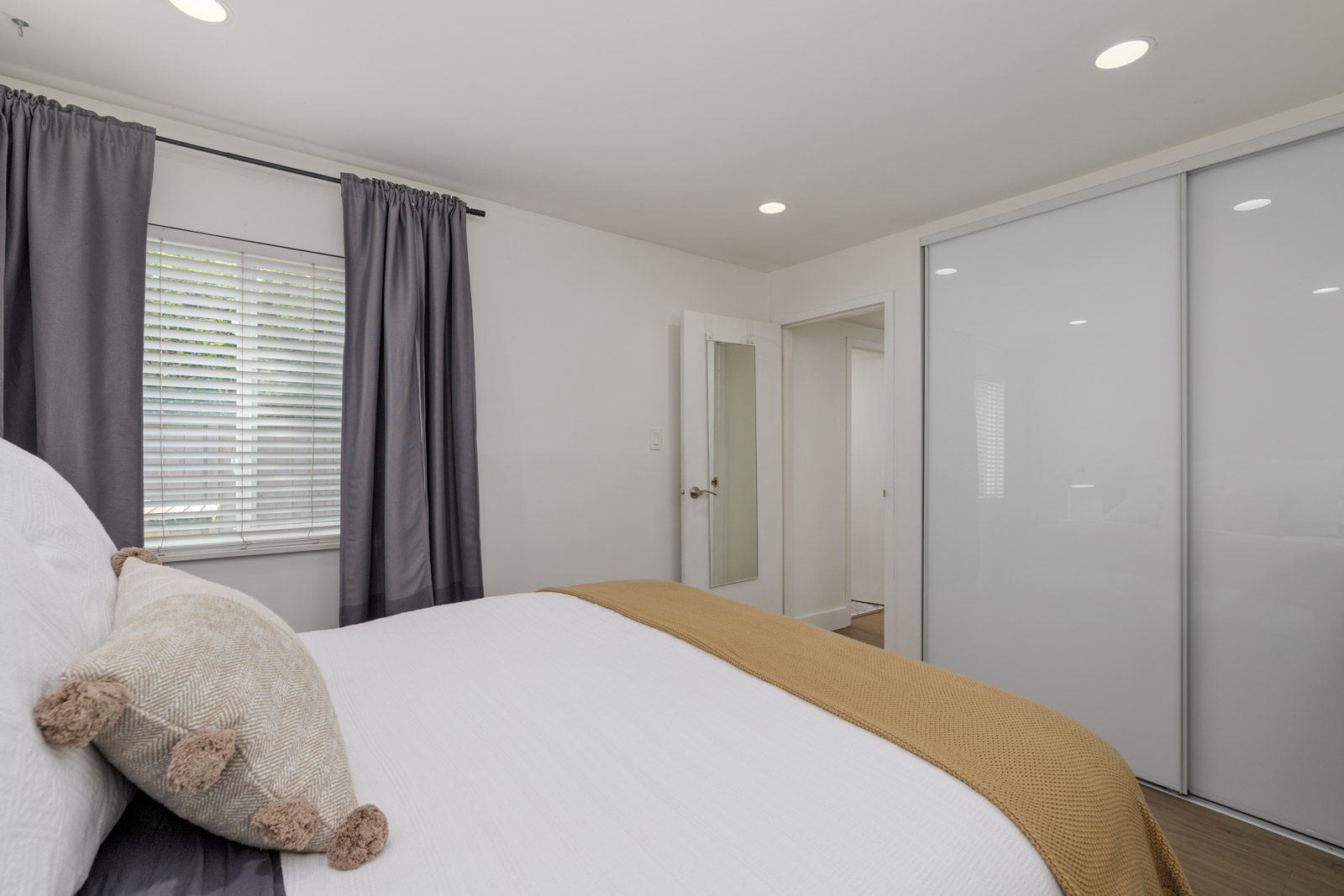 A neatly made bed with white and beige bedding stands next to a window with gray curtains in a minimalist bedroom featuring a large closet with sliding doors.