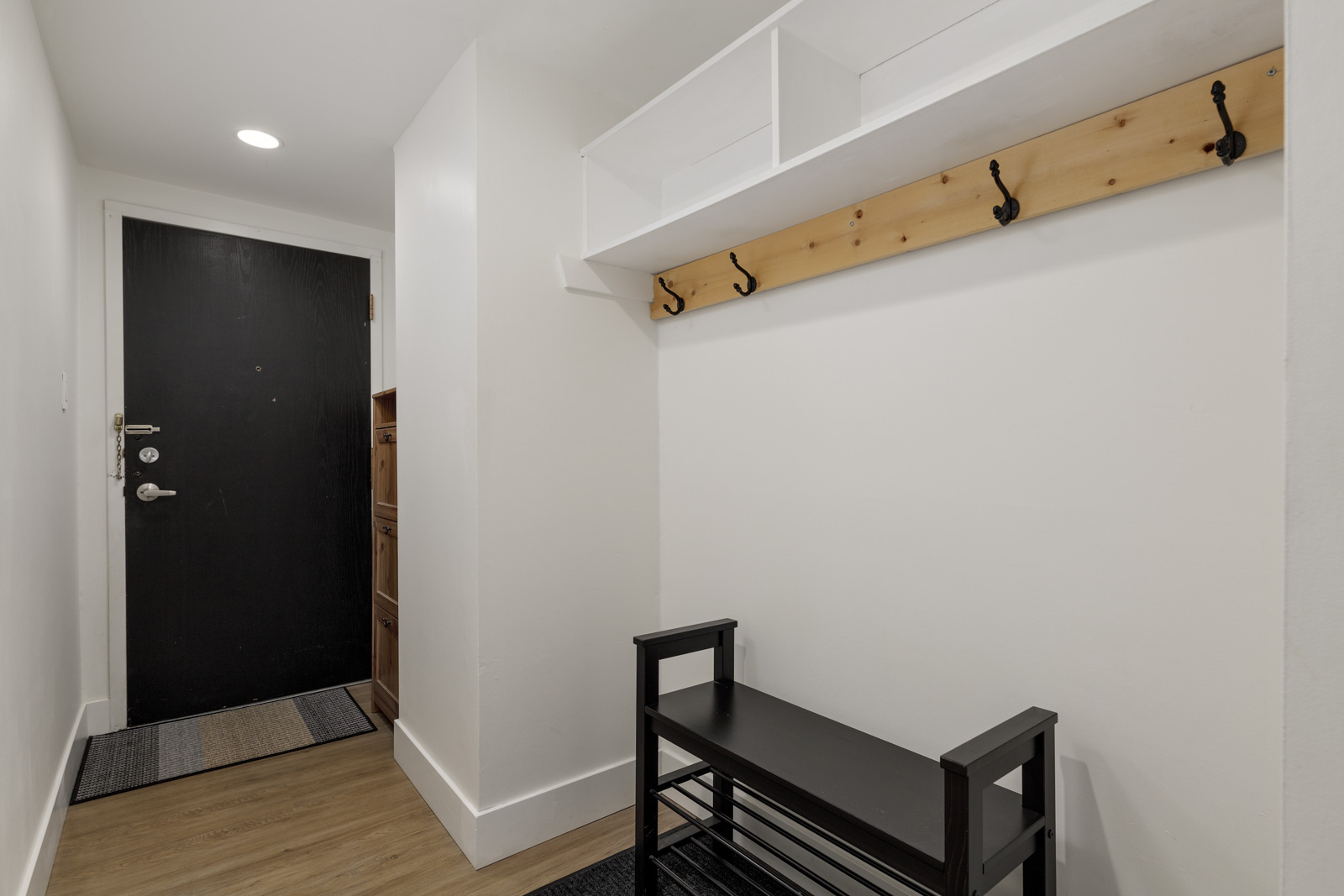 Minimalist entryway with a black front door, wall hooks on a wooden strip, a shoe bench, white walls, and wood flooring.