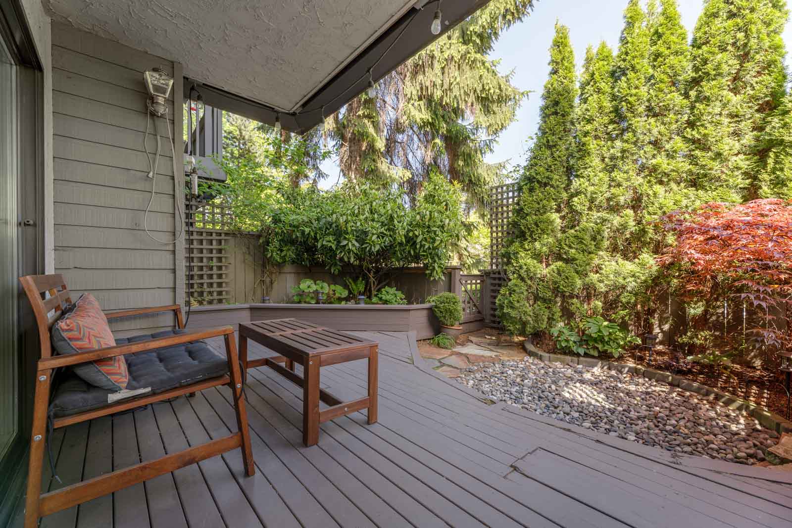 A wooden deck with two chairs and a table overlooks a small landscaped garden with gravel, shrubs, and trees surrounded by a privacy fence.