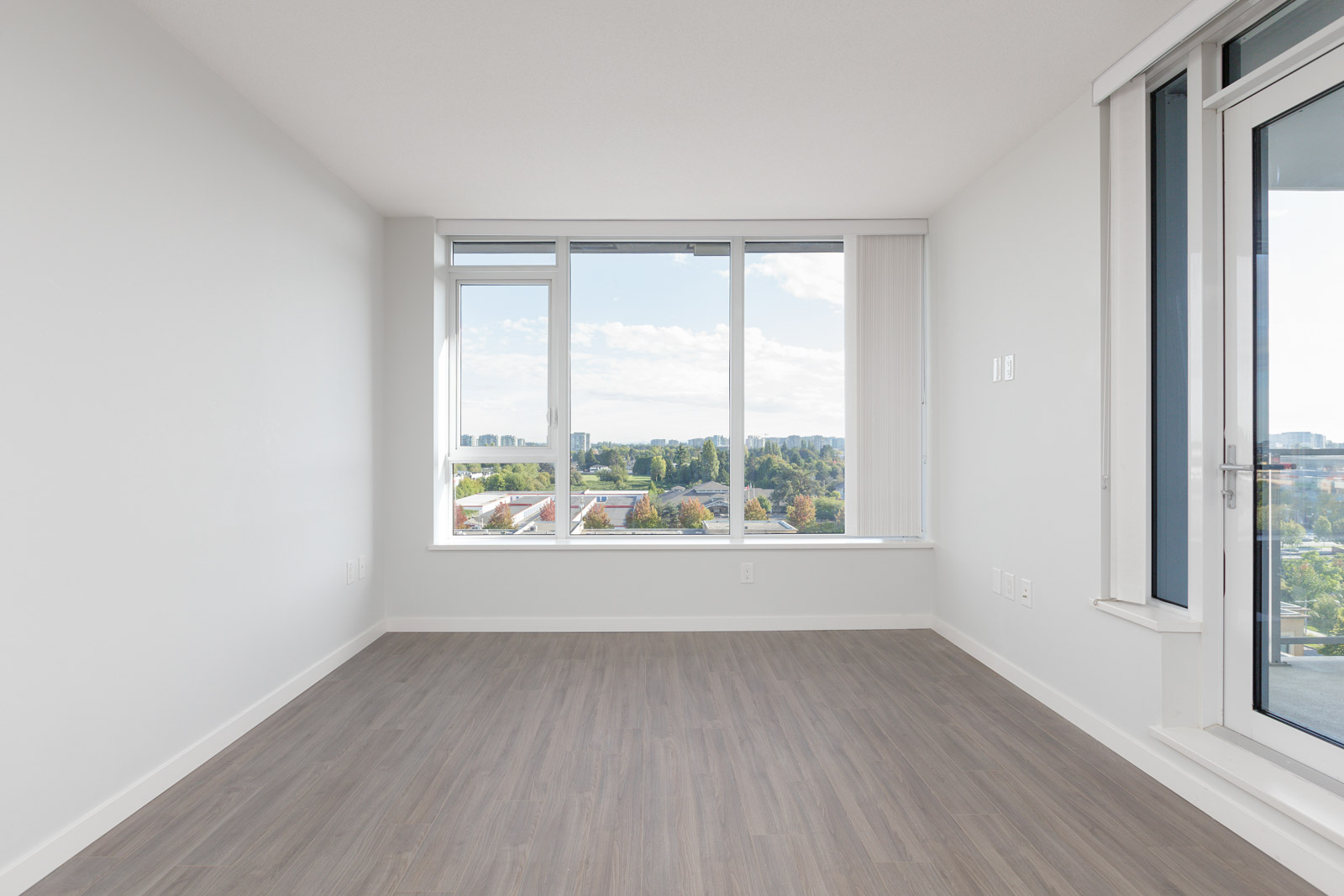 Empty modern room with large windows, light gray walls, and wood-style flooring, overlooking a cityscape and greenery outside.