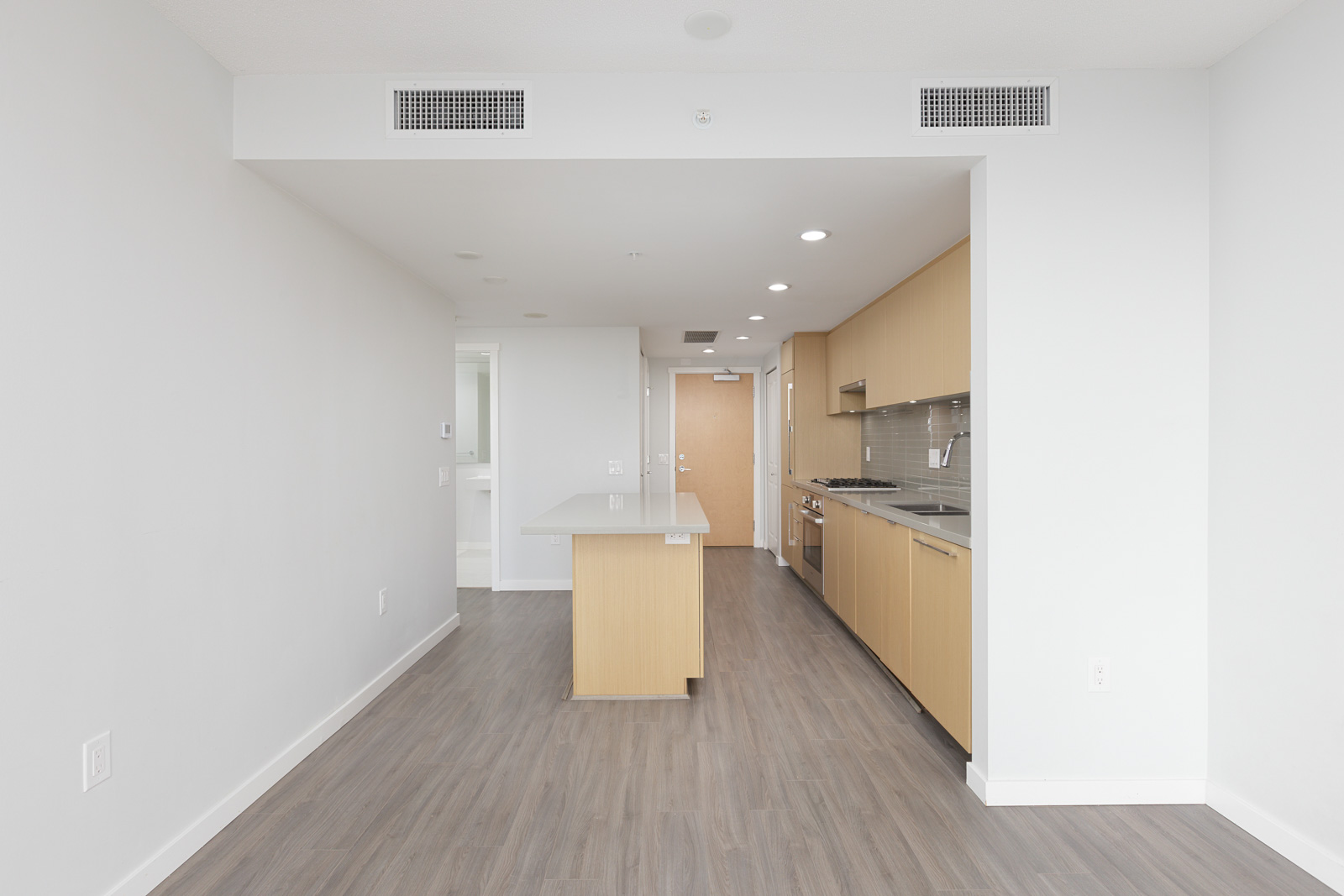 Modern, unfurnished apartment kitchen with light wood cabinets, built-in appliances, an island, and gray wood flooring. White walls and ceiling with air vents visible.