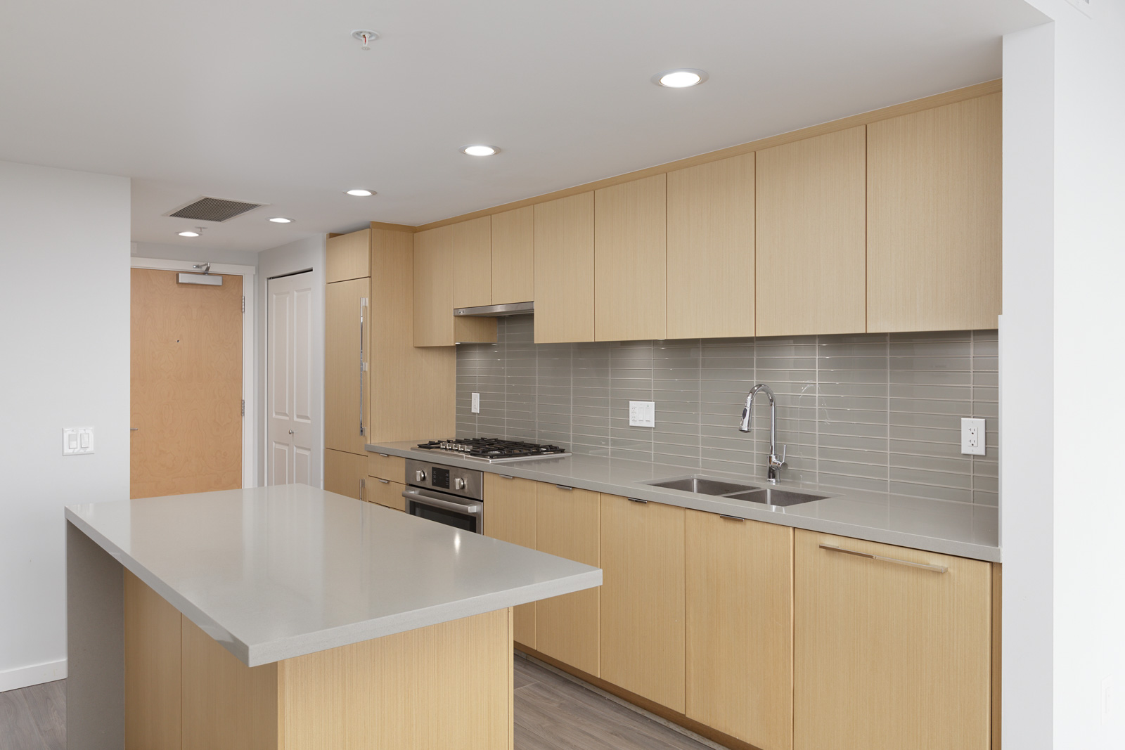 Modern kitchen with light wood cabinets, gray tile backsplash, built-in stove, sink, and a large island with a light gray countertop.