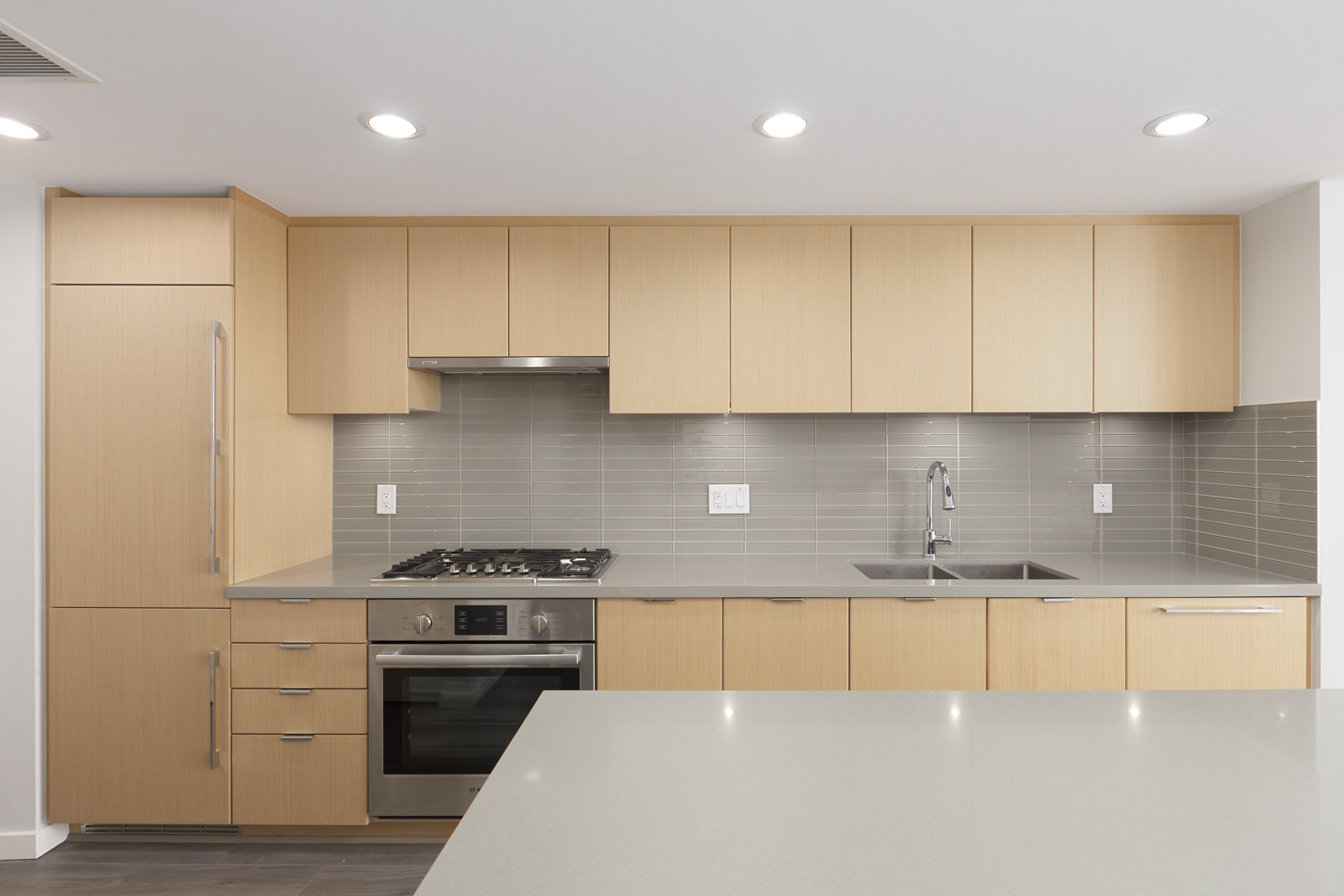 Modern kitchen with light wood cabinets, stainless steel appliances, gray tile backsplash, built-in oven, gas stove, and sink under recessed lighting.