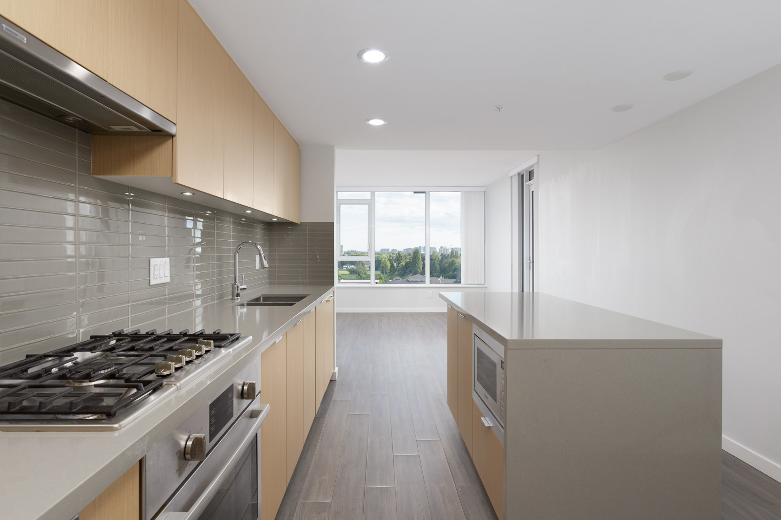 Modern kitchen with built-in gas stove, oven, microwave, and sink. Light wood cabinets, gray tile backsplash, large countertop, and window view in background.