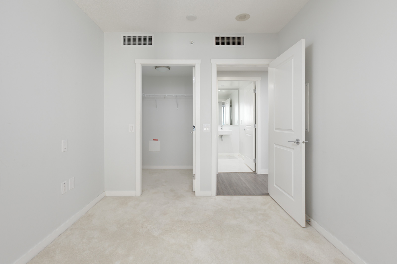 Empty white bedroom with open closet and door leading to a bathroom with white fixtures and a gray floor.