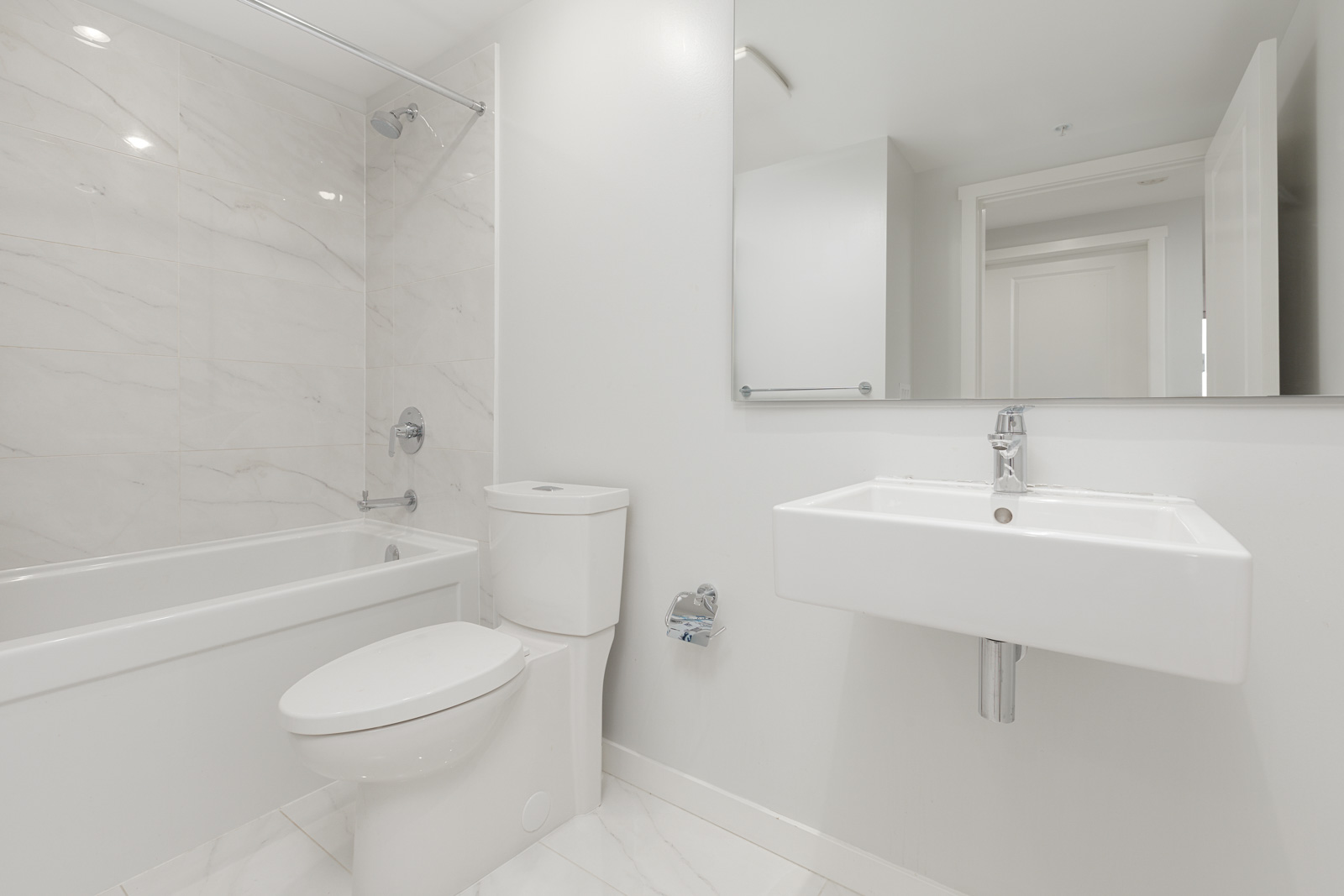 Modern bathroom with white walls, bathtub-shower combo, toilet, wall-mounted sink, large mirror, and tiled floor.