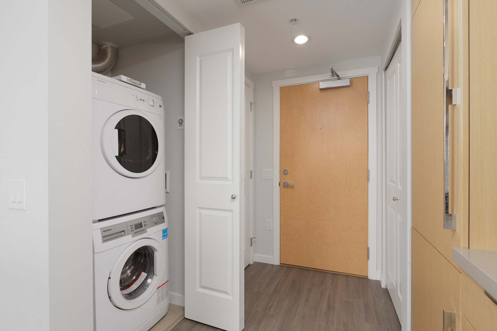 A stacked washer and dryer are in a small laundry closet with an open door next to a wooden entrance door in a modern apartment.