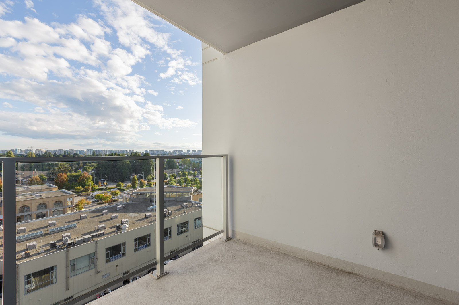 A small, empty balcony with a glass railing overlooks a cityscape with buildings, trees, and a partly cloudy sky.