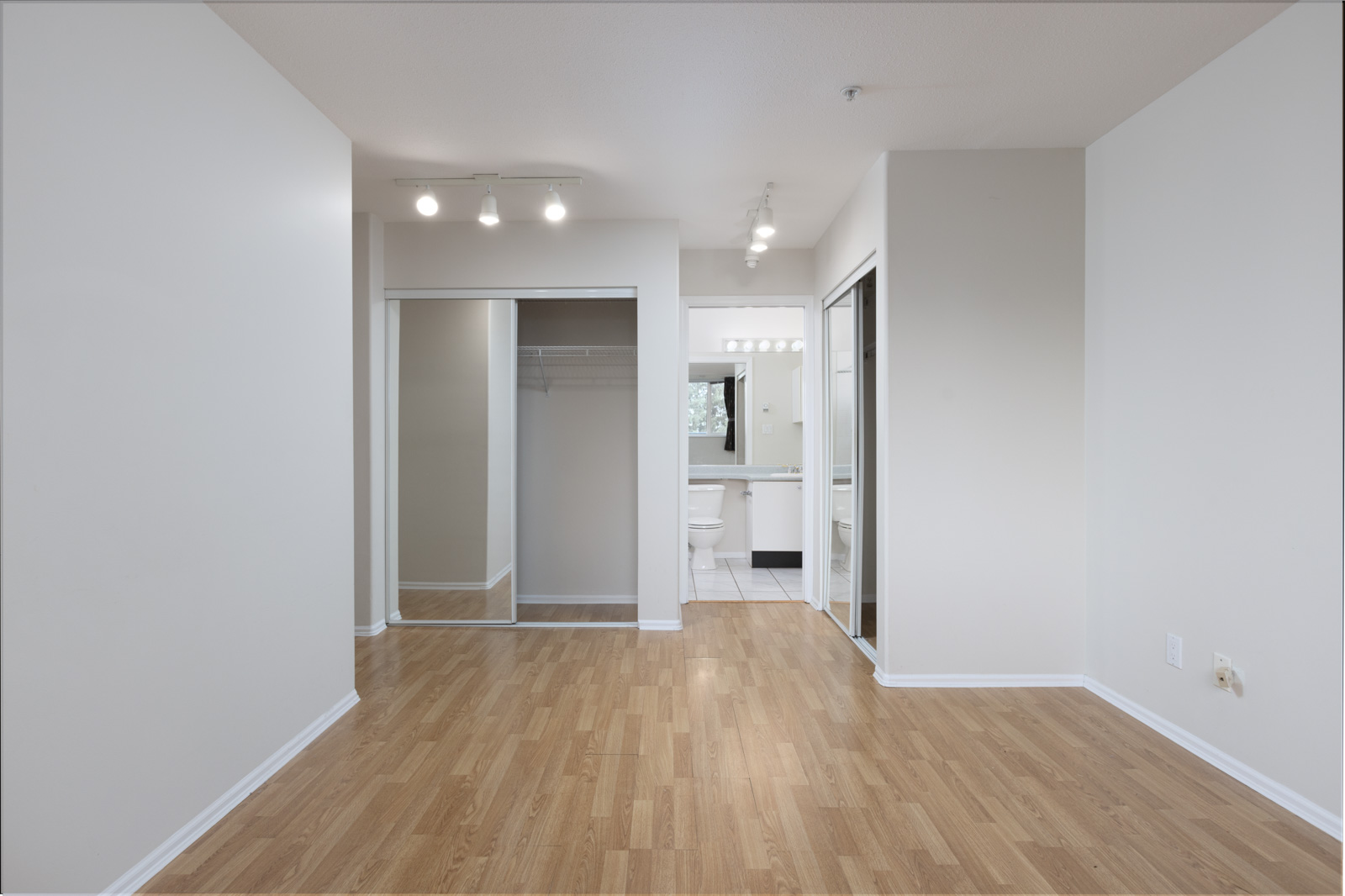 Empty room with light wood flooring, white walls, mirrored closet doors, and an attached bathroom visible in the background.