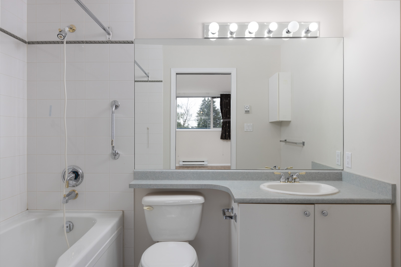 A clean, modern bathroom with a bathtub, toilet, large mirror, sink, and overhead lighting, reflected in the mirror is a window showing trees outside.