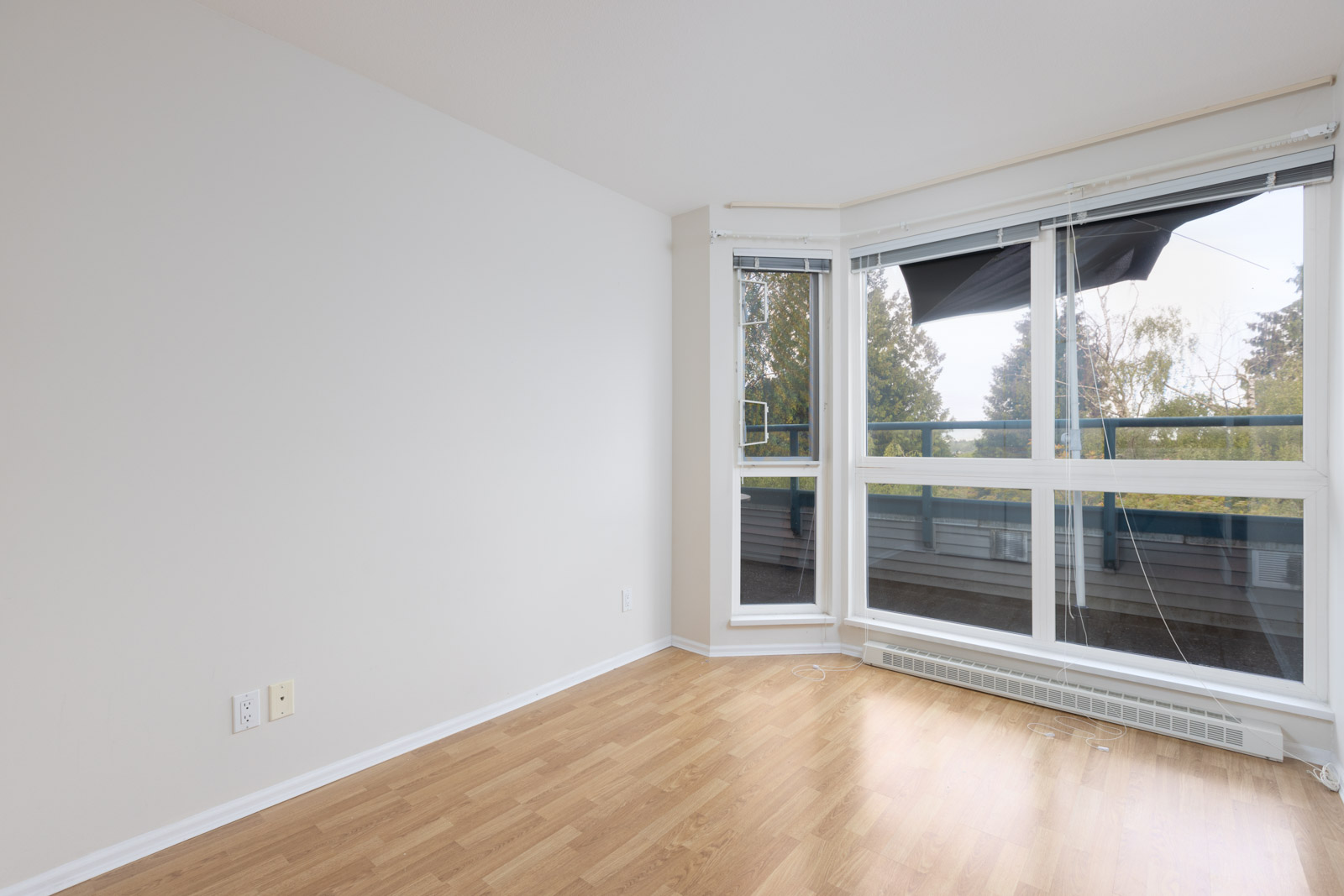 Empty room with light hardwood flooring, white walls, large corner windows, and a baseboard heater; trees and sky visible outside.