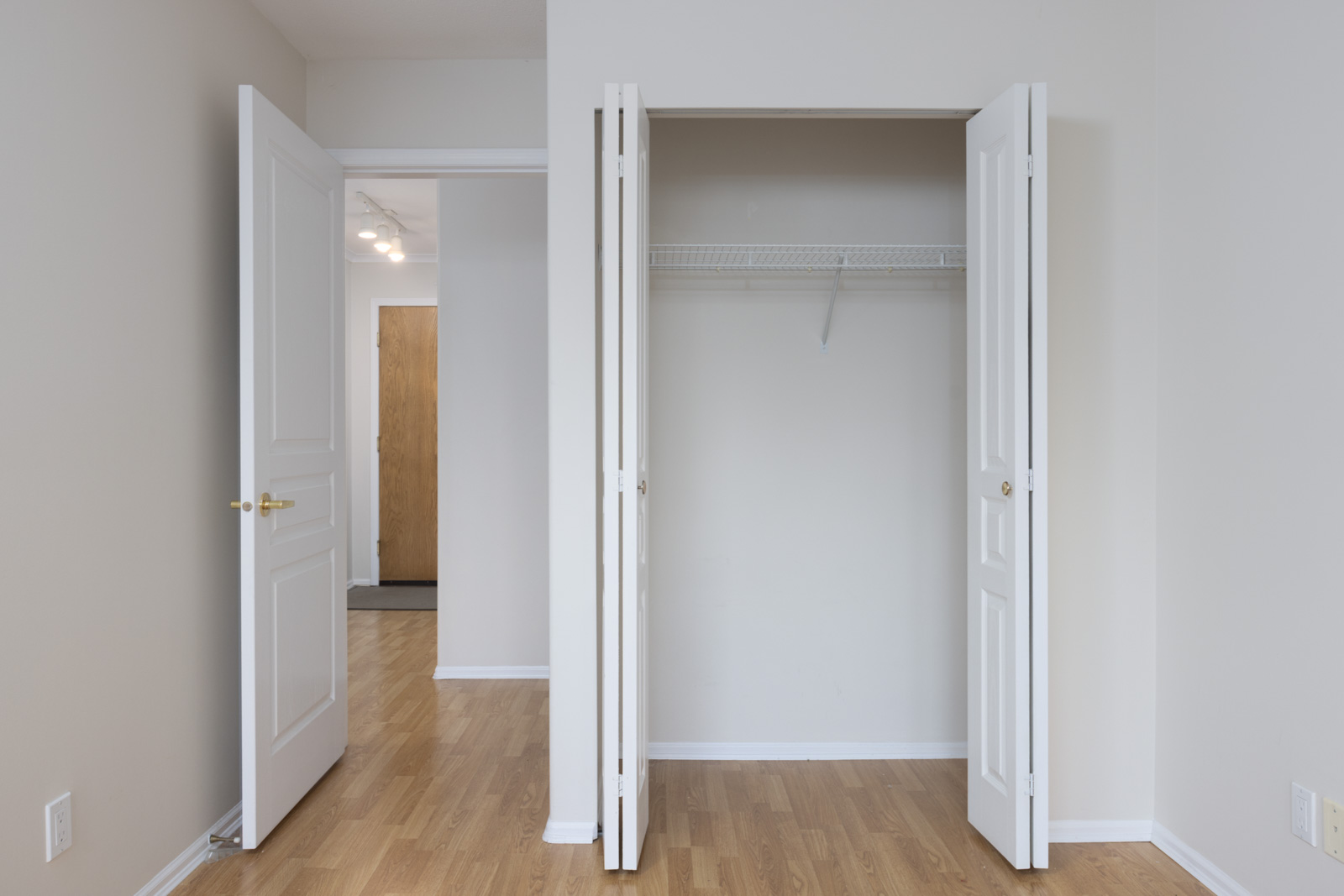 Empty room with light wood flooring, an open closet with white bi-fold doors, and an open white door leading to a hallway. Walls are painted off-white.