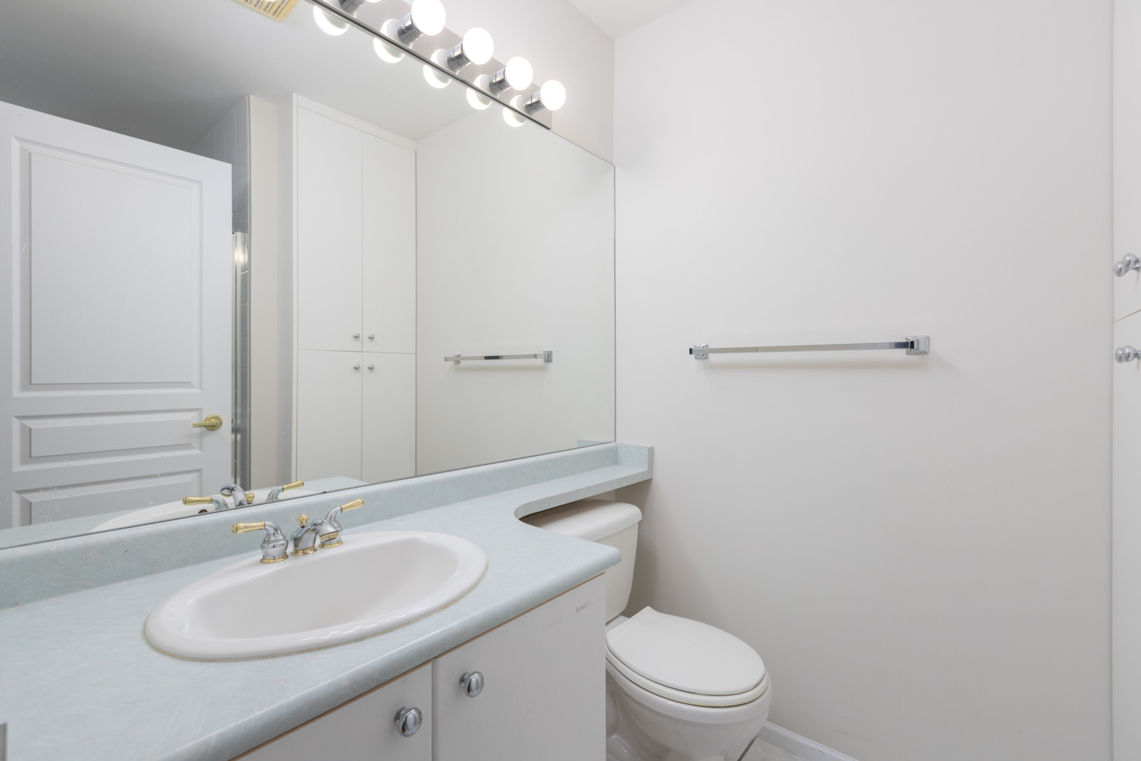 A modern bathroom with a sink, countertop, toilet, large mirror, wall-mounted lights, towel bar, and white cabinets.
