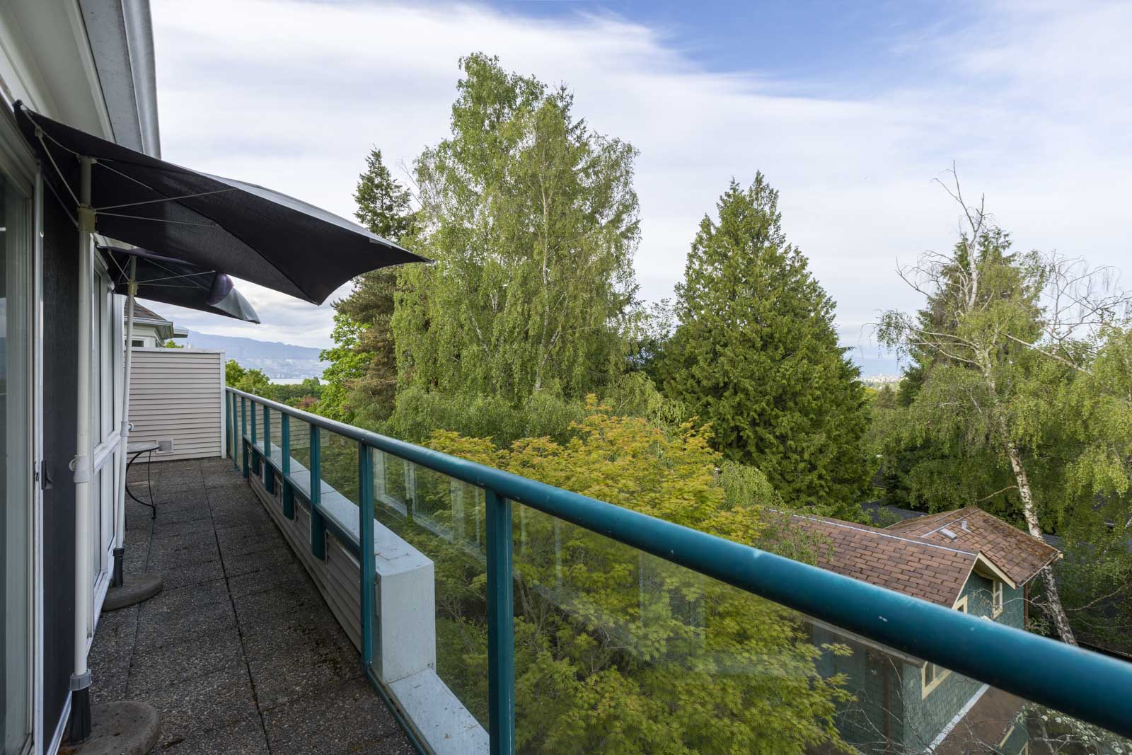 A balcony with glass railing offers a view of trees and neighboring house under a partly cloudy sky.