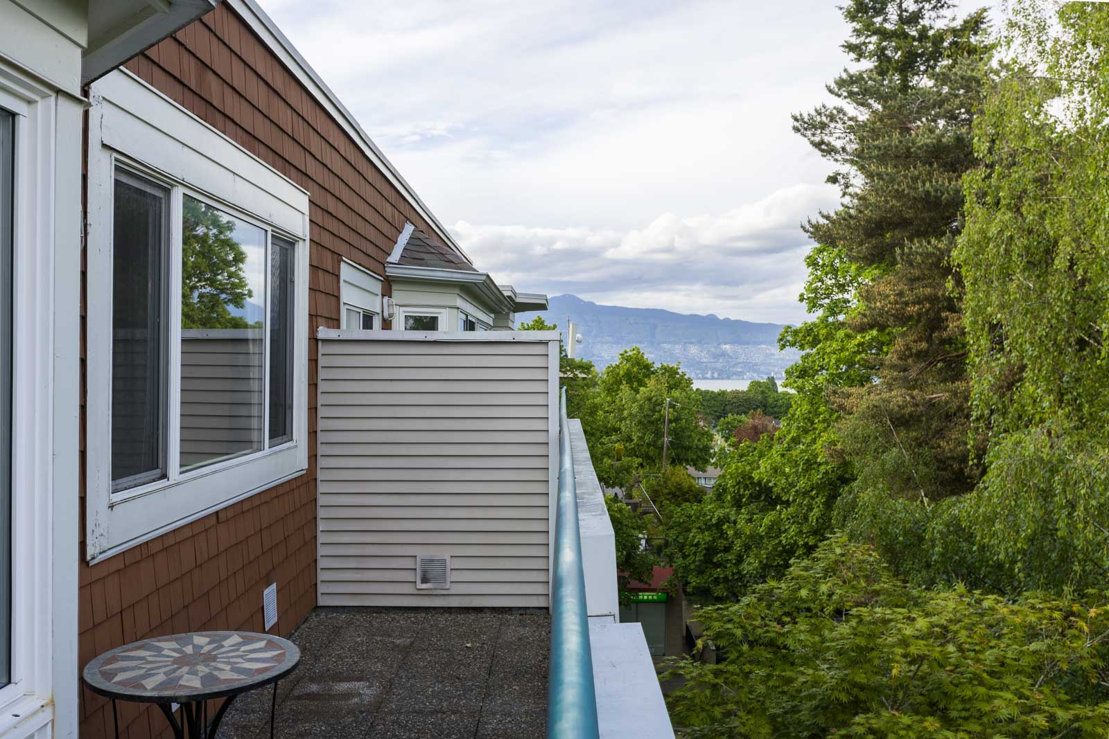 A small balcony with a round table overlooks green trees and distant mountains under a cloudy sky.