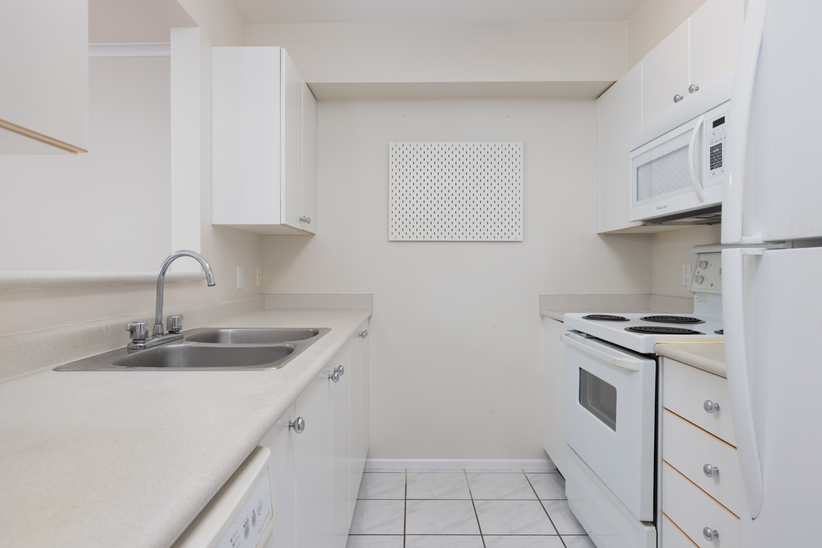 A small, white kitchen with a double sink, electric stove, microwave, and refrigerator, featuring white cabinets and tile flooring.