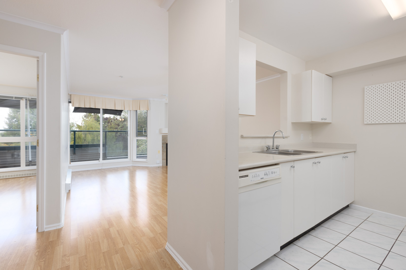 Open-plan apartment with a white kitchen on tiled flooring, adjacent to a living area with wood flooring, large windows, and a balcony view.