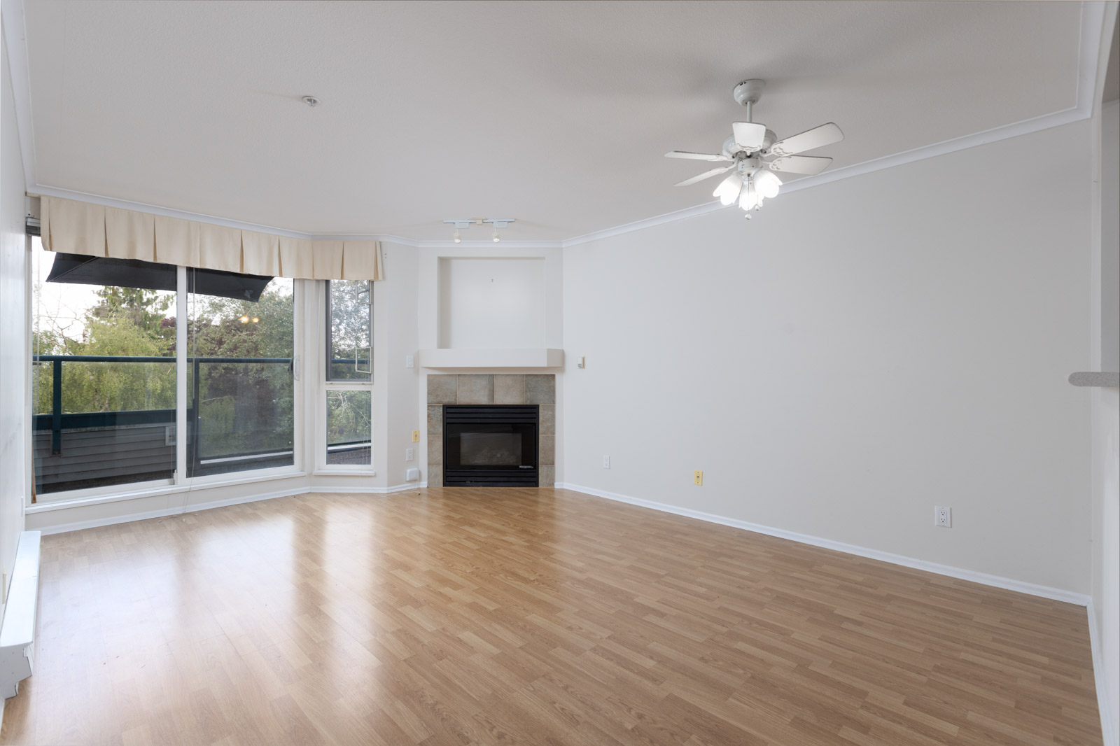Empty living room with light wood flooring, white walls, a ceiling fan with lights, fireplace, and large windows overlooking a balcony with trees visible outside.
