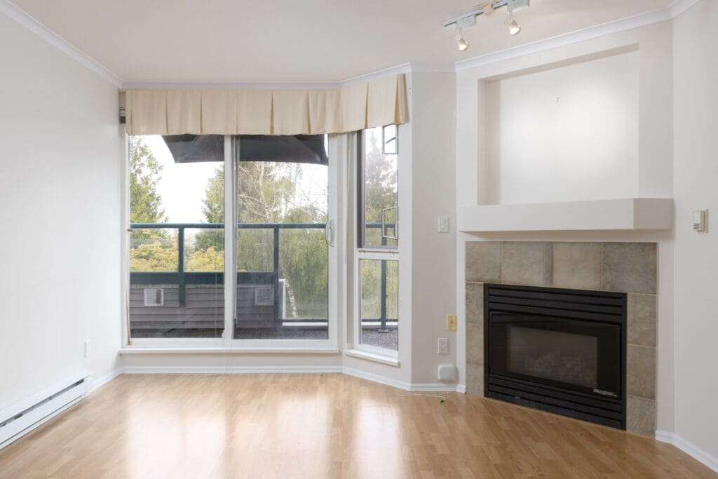 Unfurnished living room with light wood flooring, a gas fireplace with tile surround, large windows, and a partial view of a balcony and trees outside.