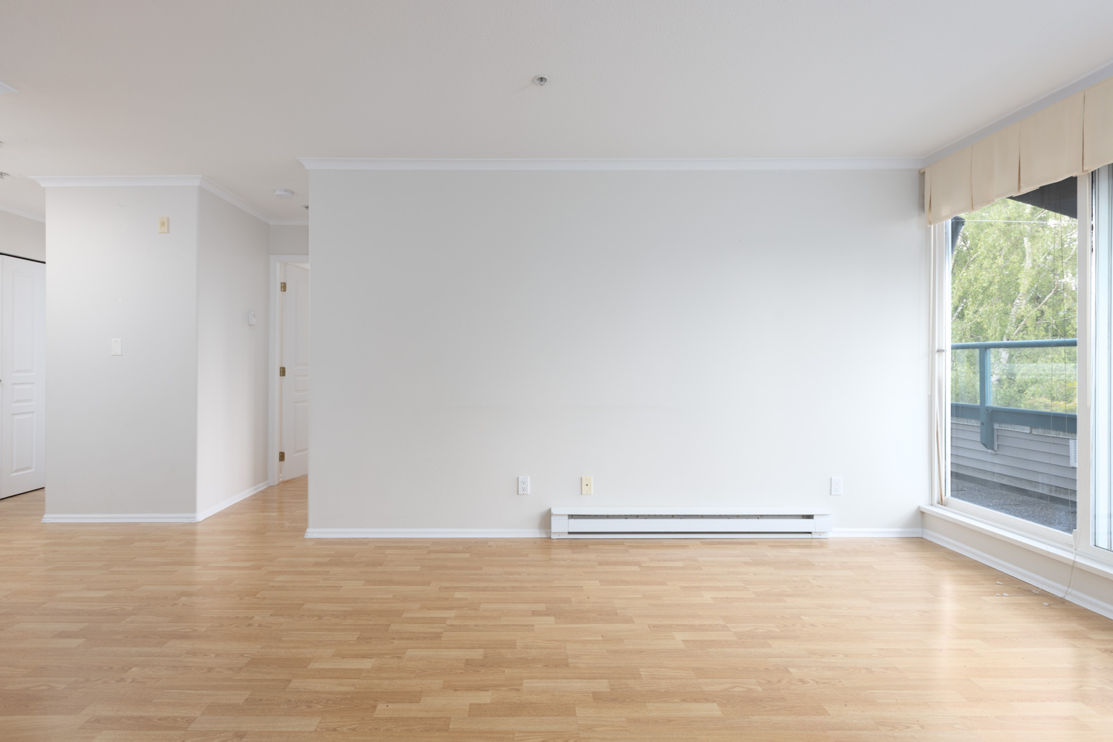 Empty room with light wood flooring, white walls, baseboard heater, large window on the right, and doors visible in the background.