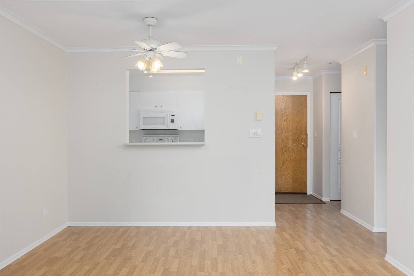 An empty room with light wood flooring, white walls, a ceiling fan, and a small kitchen area visible through a serving hatch. A wooden entrance door is in the background.