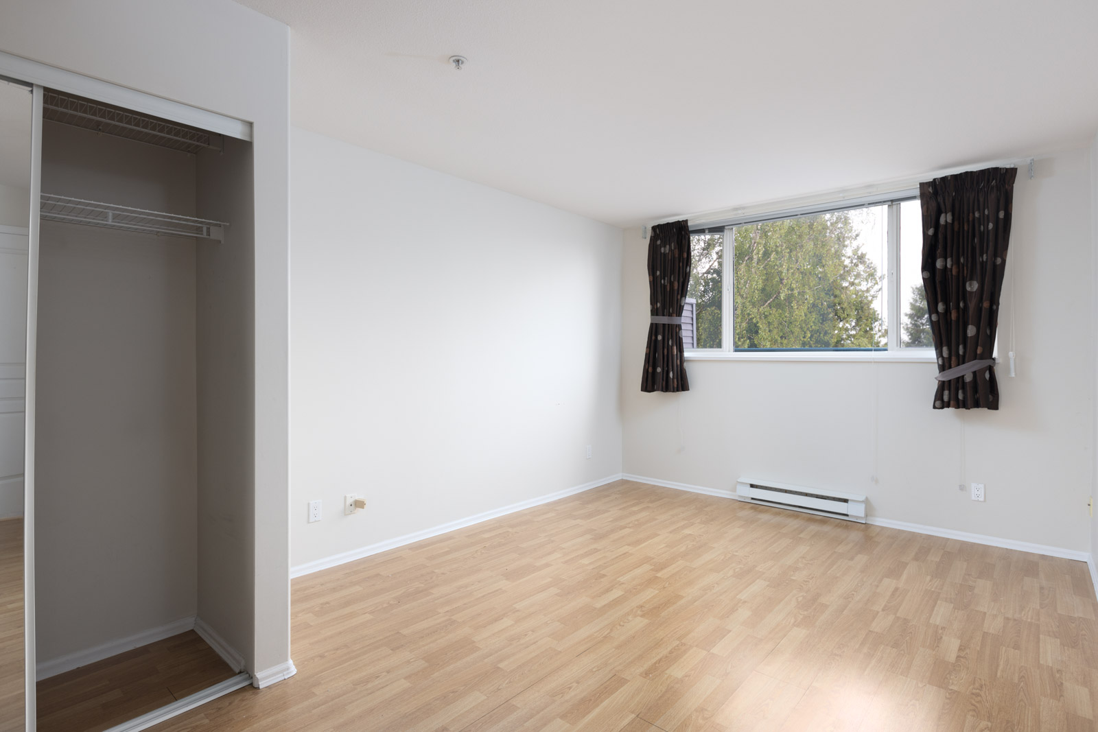 Empty room with light wood flooring, a window with dark curtains, a baseboard heater, and an open closet with sliding mirrored doors.