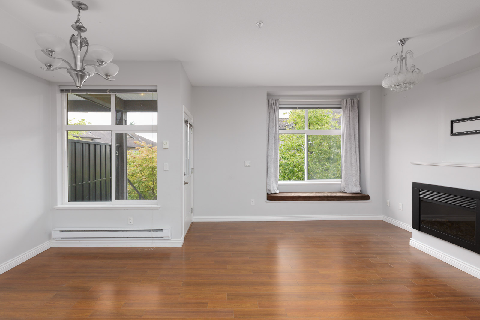 Empty living room with wood flooring, large windows, a fireplace, and two ceiling light fixtures. Natural light coming in from the windows highlights the clean, minimalist space.