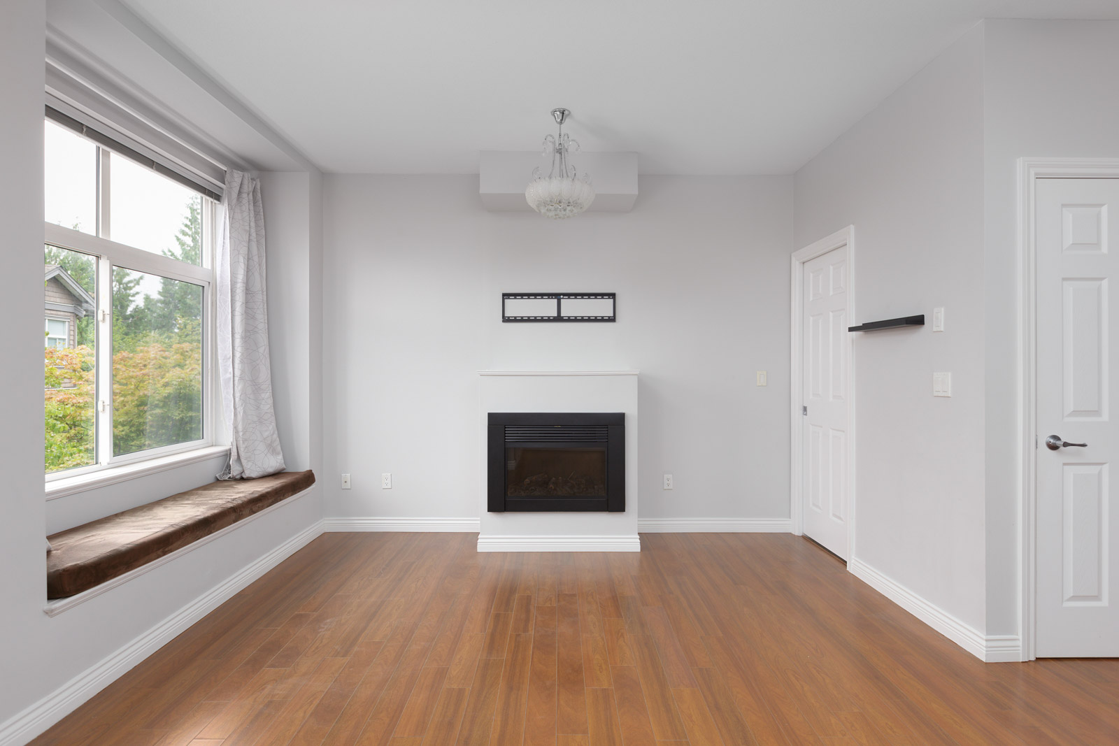 Bright, empty living room with a large window seat, light gray walls, a central fireplace, wood flooring, and a white ceiling light fixture.