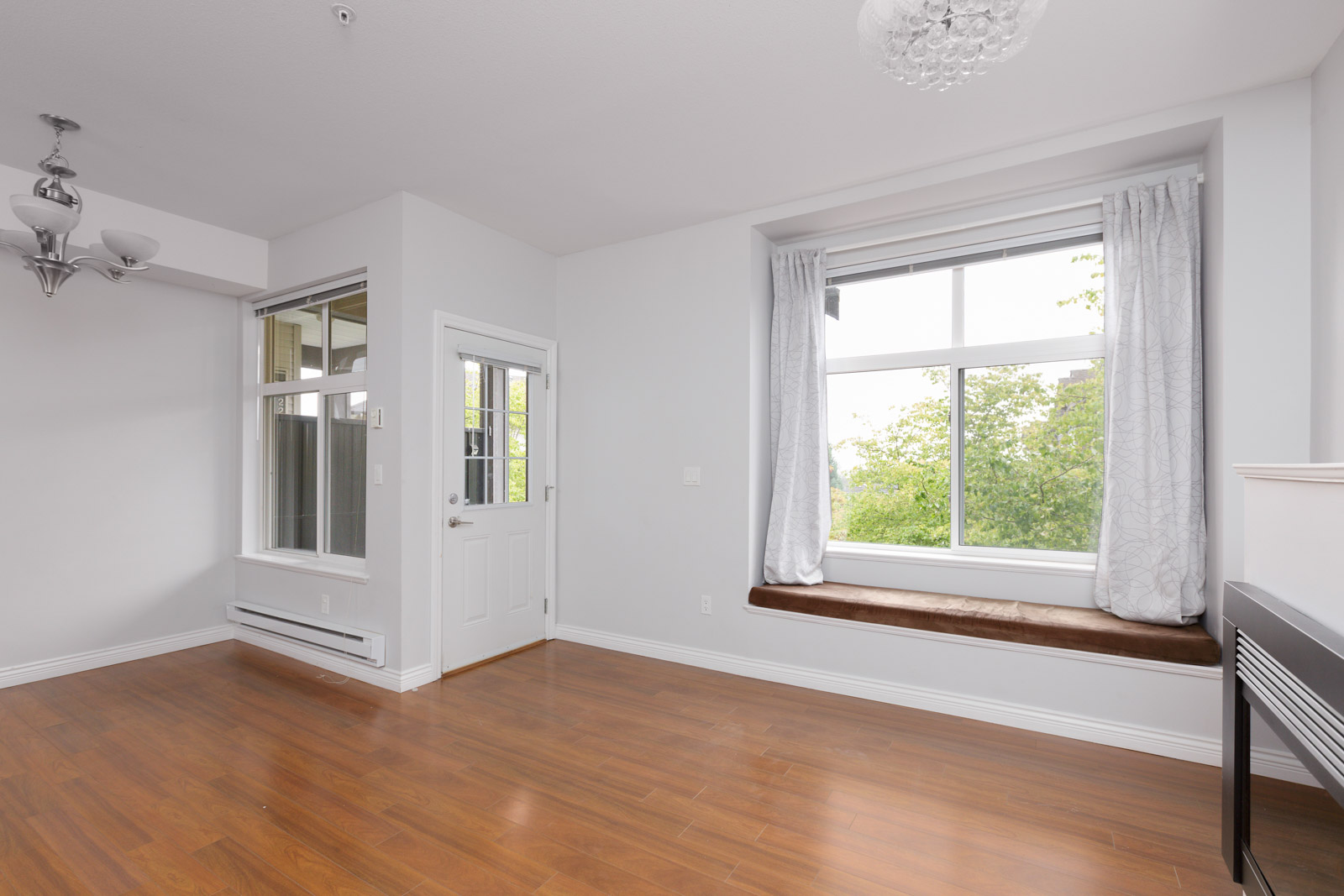 Empty room with hardwood floor, large window with white curtains and a brown window seat, white walls, and a white door leading outside.
