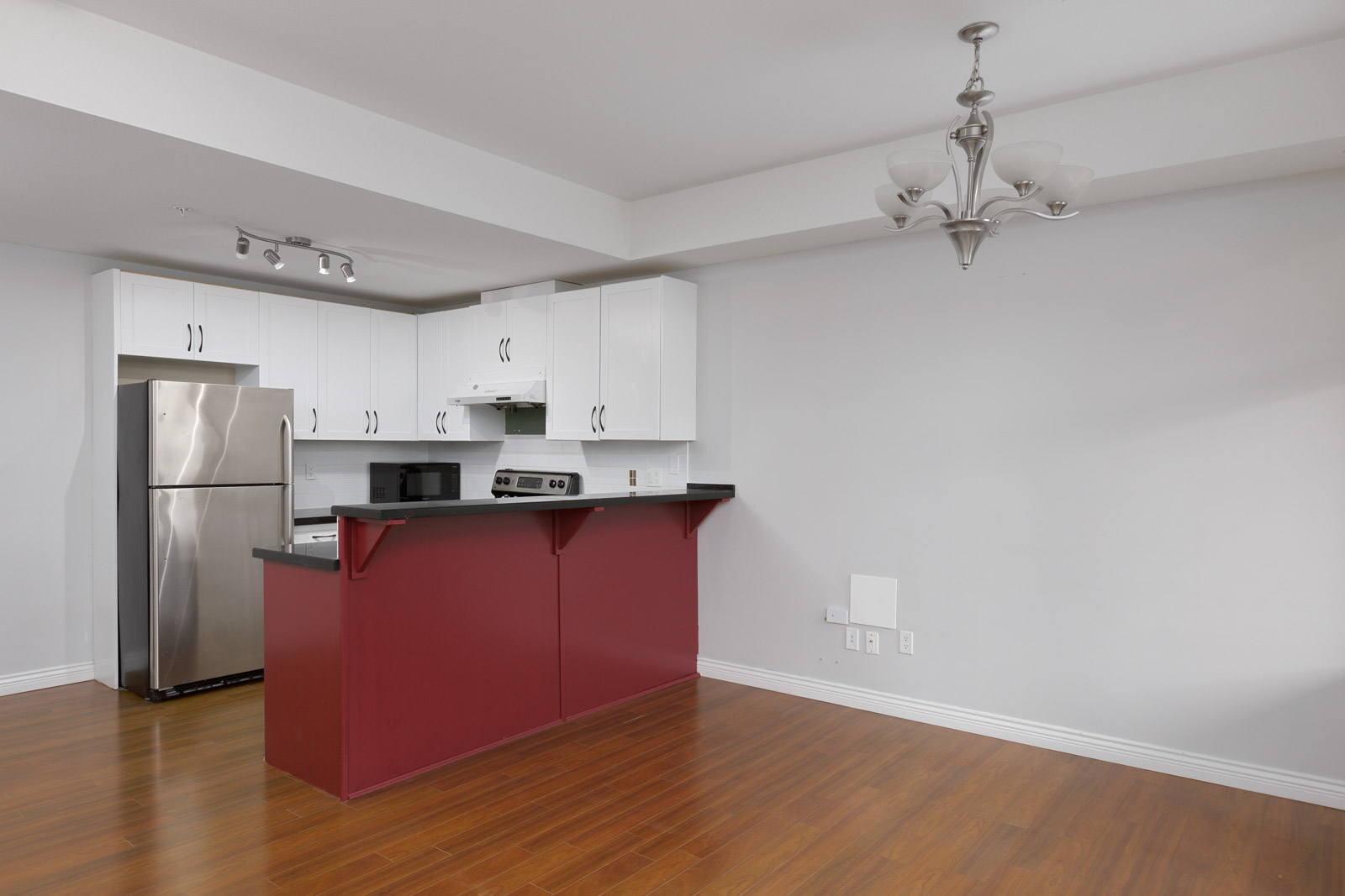Modern kitchen with stainless steel appliances, white cabinets, a red island, wood flooring, and a ceiling light fixture in an empty open-plan space.
