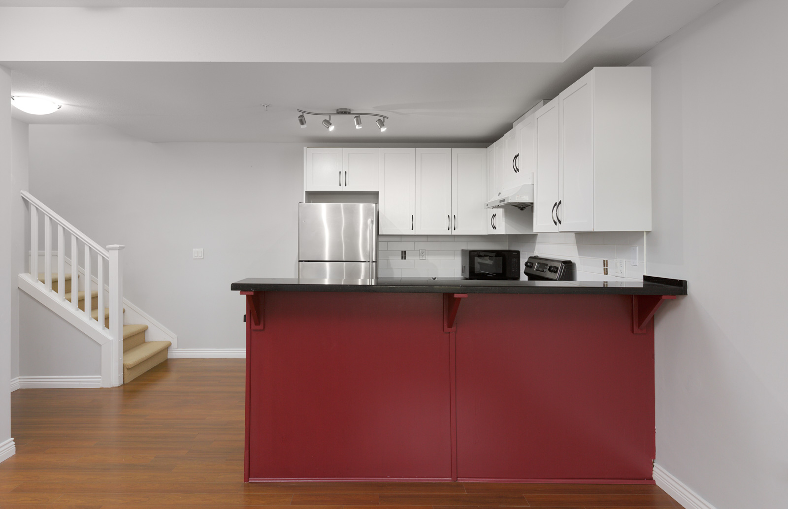 Modern kitchen with white cabinets, stainless steel appliances, red island counter, wooden floor, and staircase to the left.