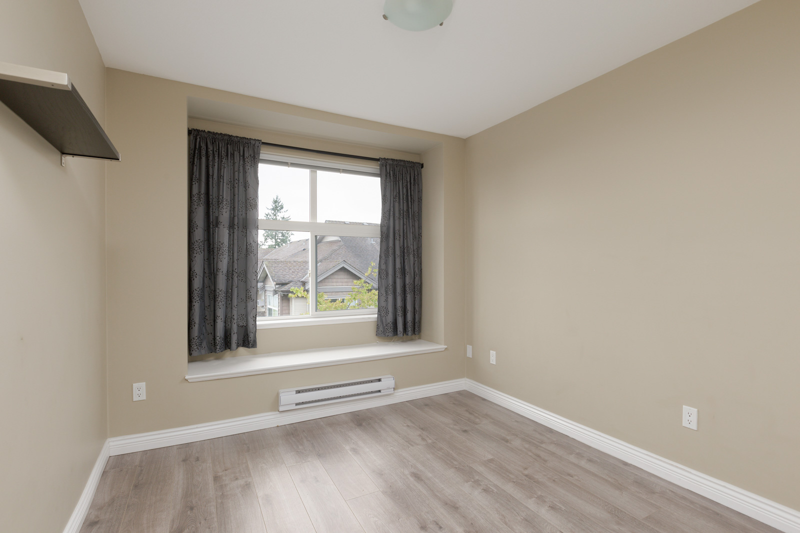Empty room with beige walls, light wood flooring, a window with dark curtains, white trim, and a baseboard heater under the window.