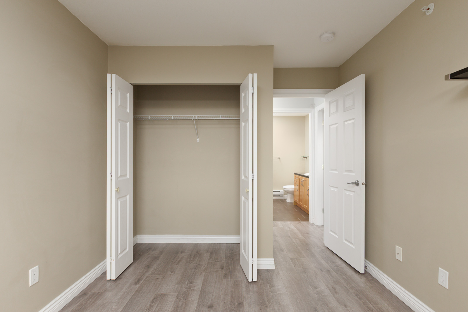 Empty beige bedroom with open double closet doors and an open door leading to a bathroom with wood cabinetry and a toilet in view.