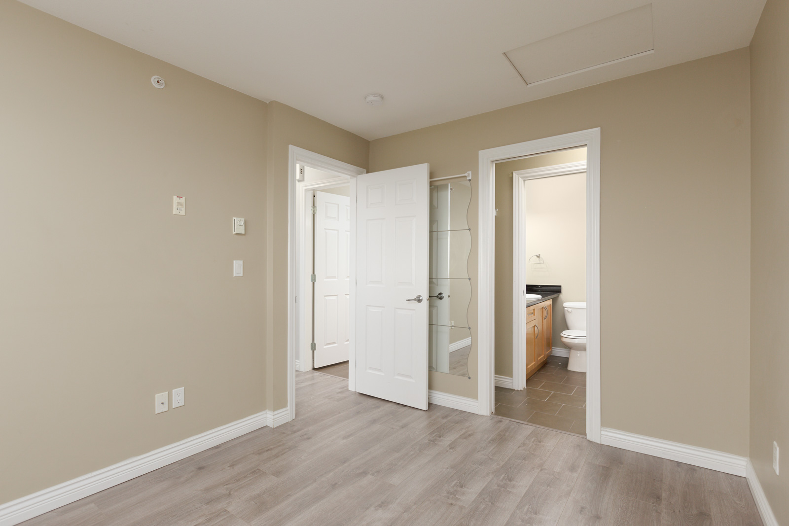 Empty room with beige walls and light wood flooring, open door leading to a bathroom with a visible toilet and vanity.