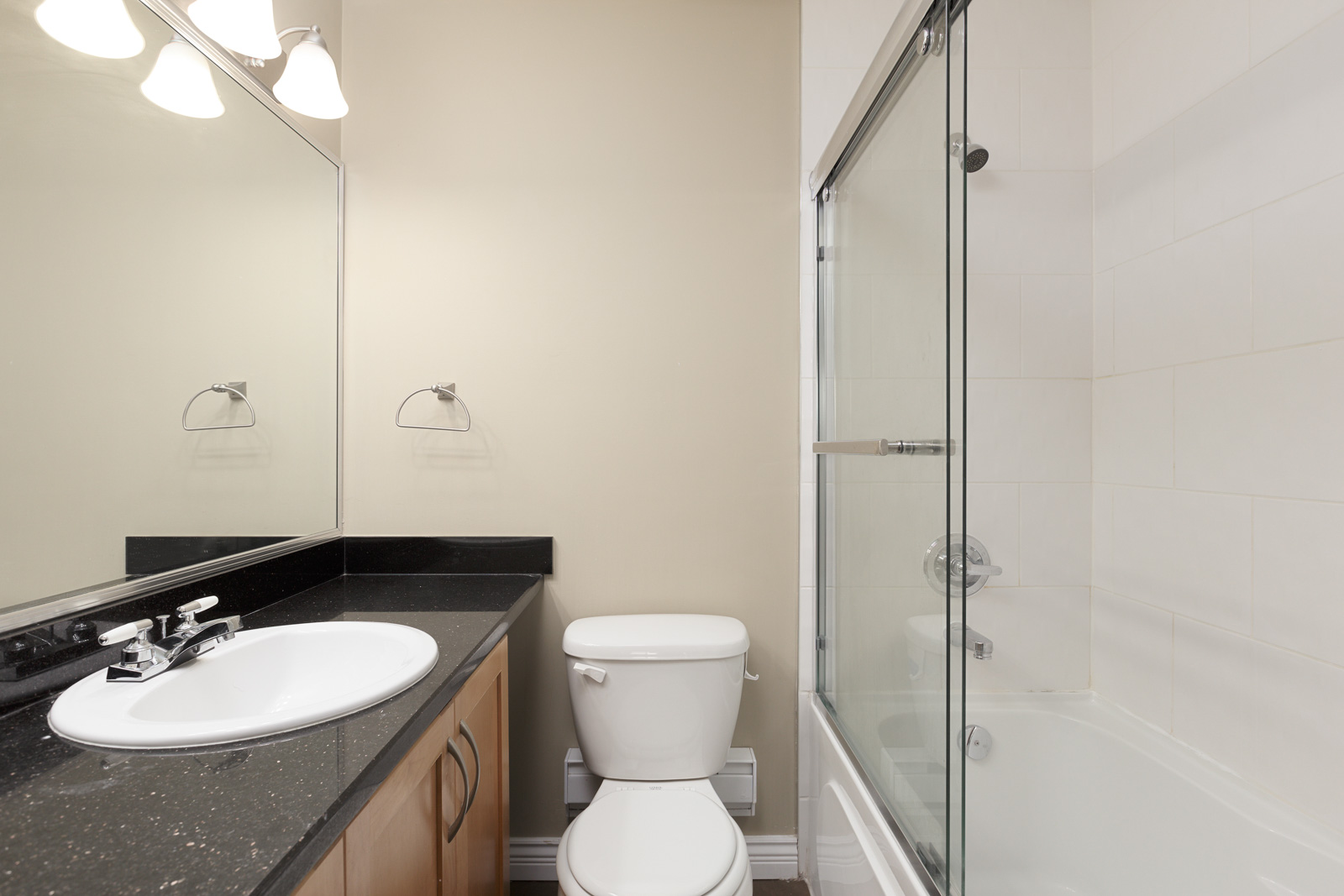 A modern bathroom with a black countertop sink, a toilet, and a bathtub with a glass shower door; neutral wall colors and overhead lighting.