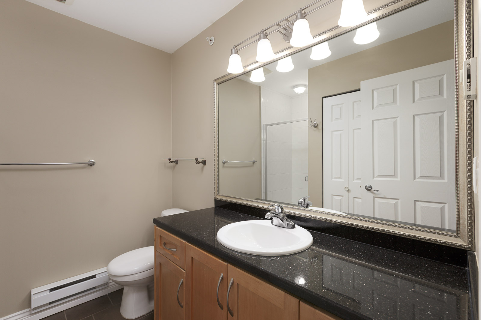 Modern bathroom with beige walls, large mirror, black countertop with single sink, wooden cabinets, toilet, and overhead lights.