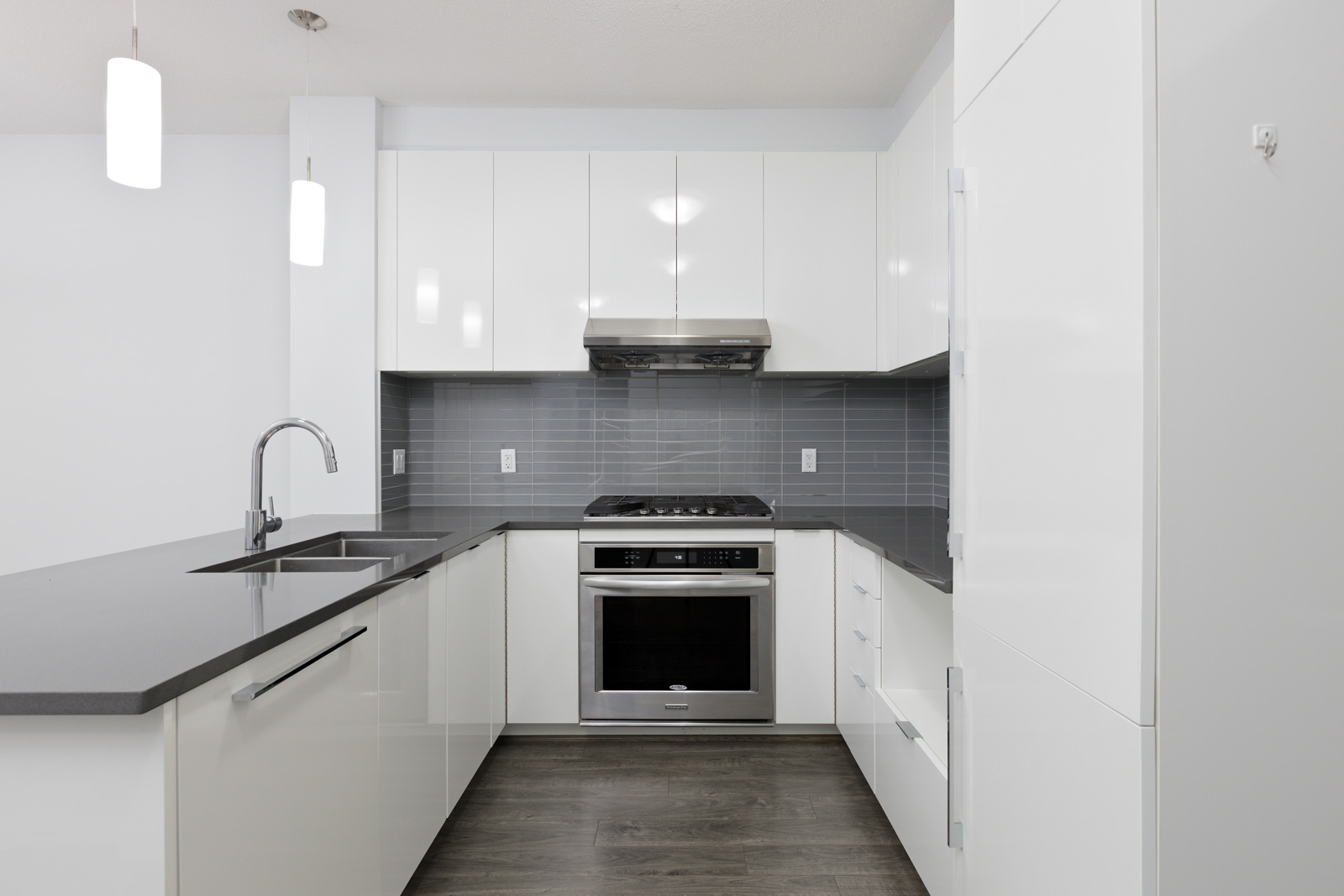 Modern kitchen with white cabinets, gray countertops, stainless steel oven, and gray tile backsplash; pendant lights hang above the sink area.
