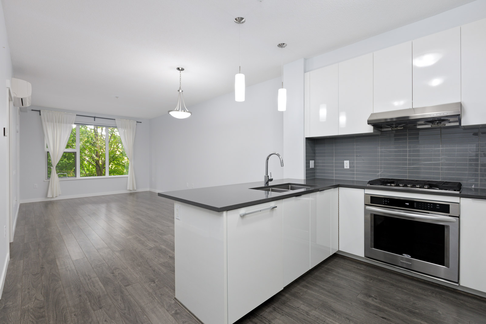Modern open-concept kitchen and living area with white cabinets, grey countertops, stainless steel appliances, light wood flooring, and large window with white curtains.