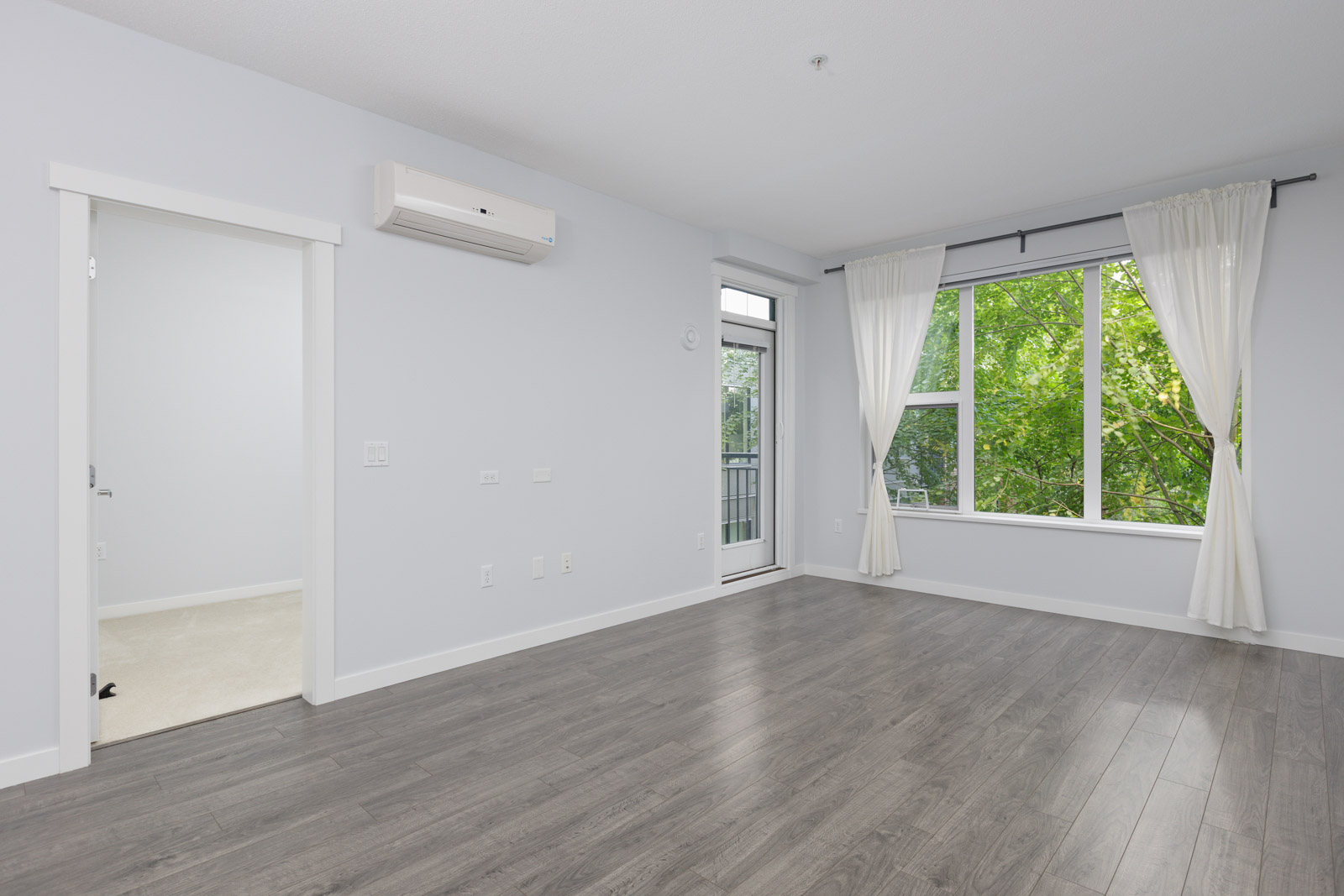 Empty room with gray wood flooring, white walls, a large window with white curtains, and a wall-mounted air conditioner. A door leads to another room and a glass door leads outside.