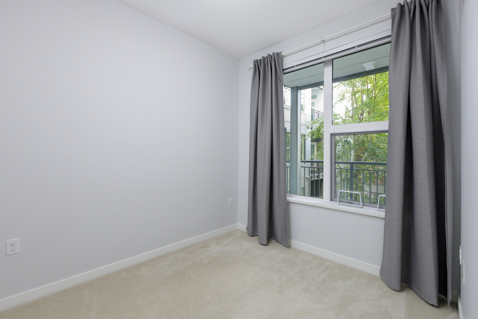Empty room with light gray walls, beige carpet, and a window with gray curtains overlooking a balcony and greenery outside.
