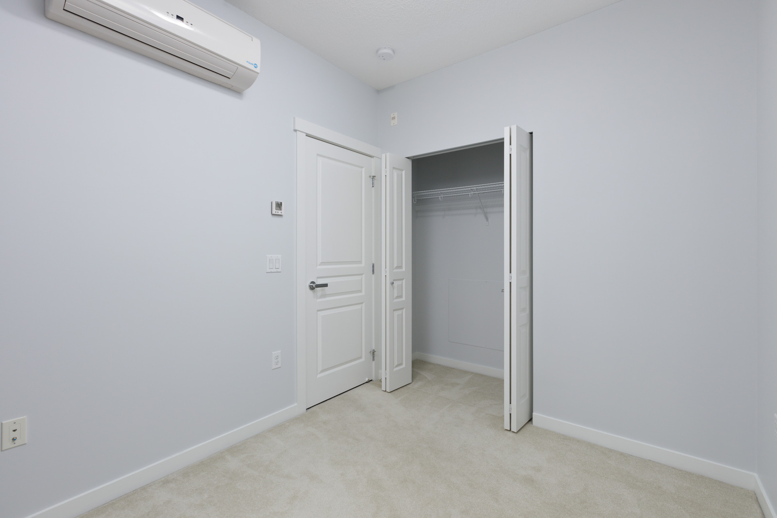 Empty room with light gray walls, beige carpet, a double door closet, and a wall-mounted air conditioning unit above a closed door.
