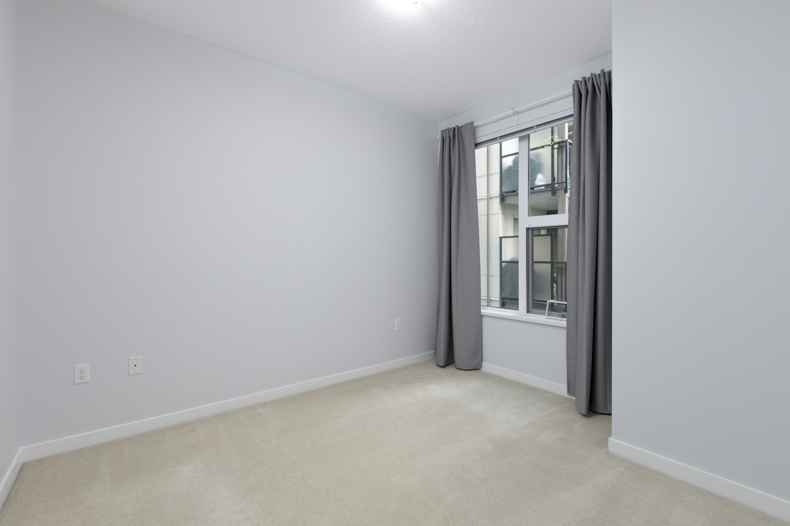 Empty room with light-colored carpet, white walls, two electrical outlets, a window with gray curtains, and natural light coming in.