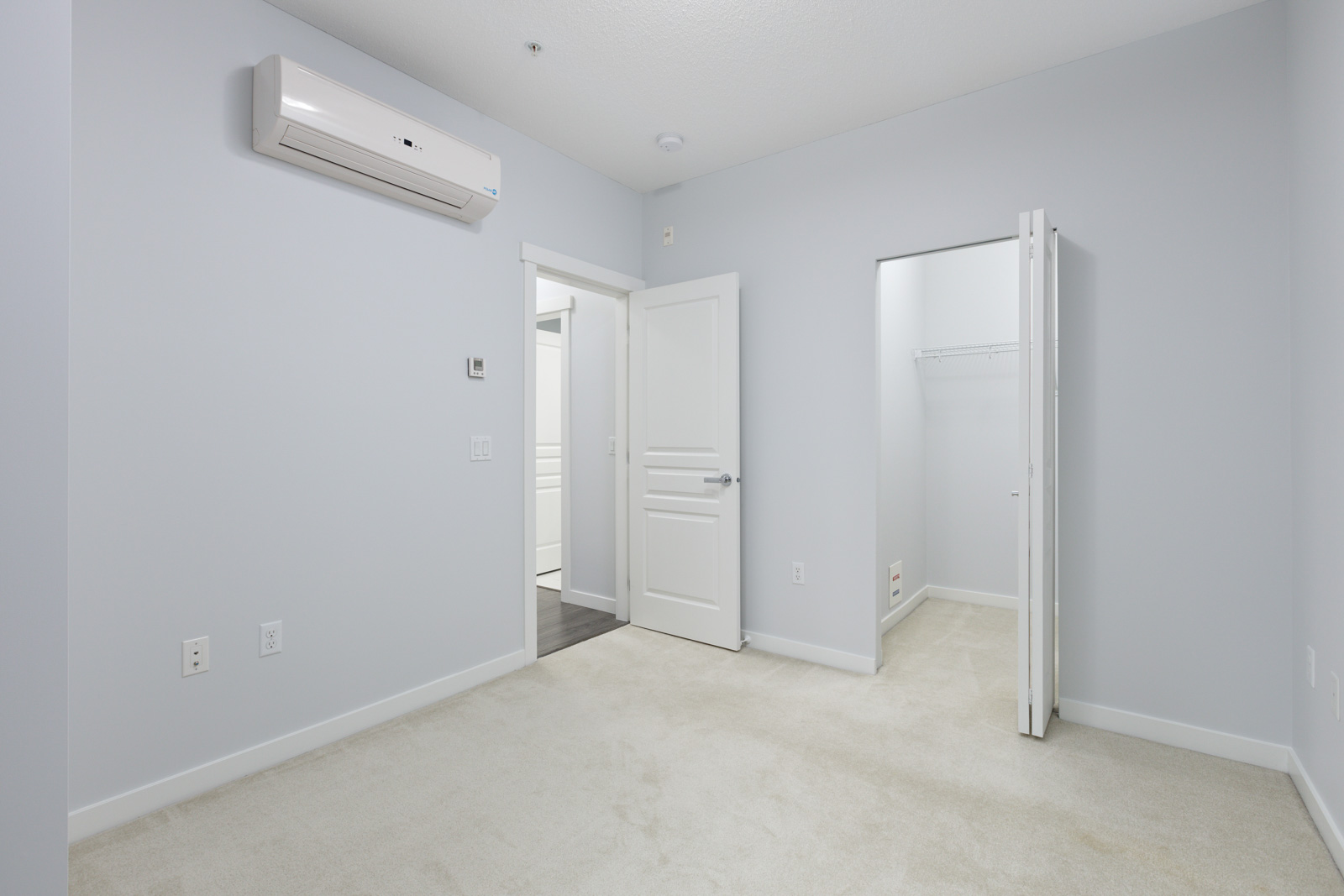 Empty room with light gray walls, beige carpet, white doors, a wall-mounted air conditioning unit, and an open closet with a shelf and hanging rod.