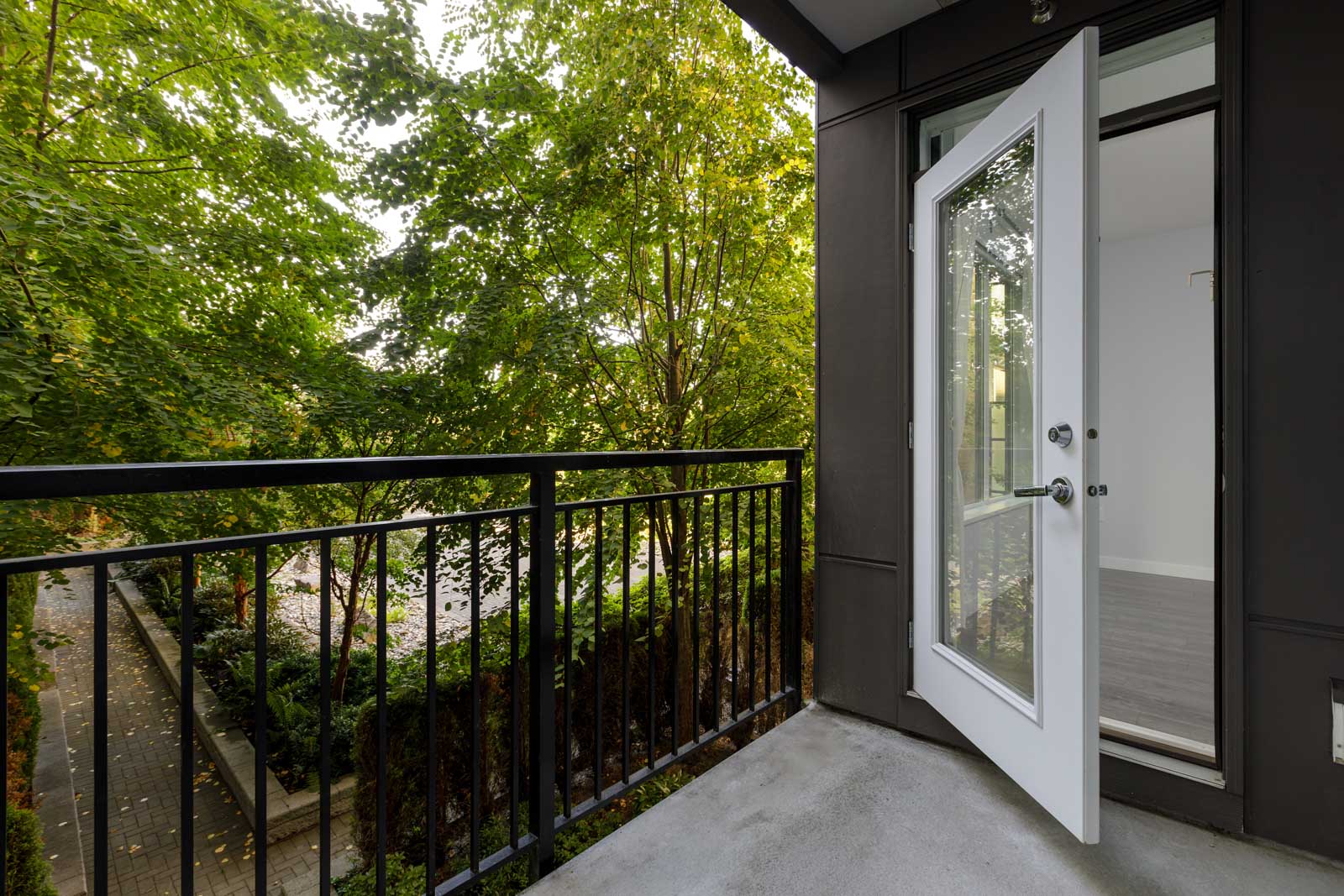 Open glass door leads from a room to a small balcony with black railing, overlooking green trees and a paved walkway below.
