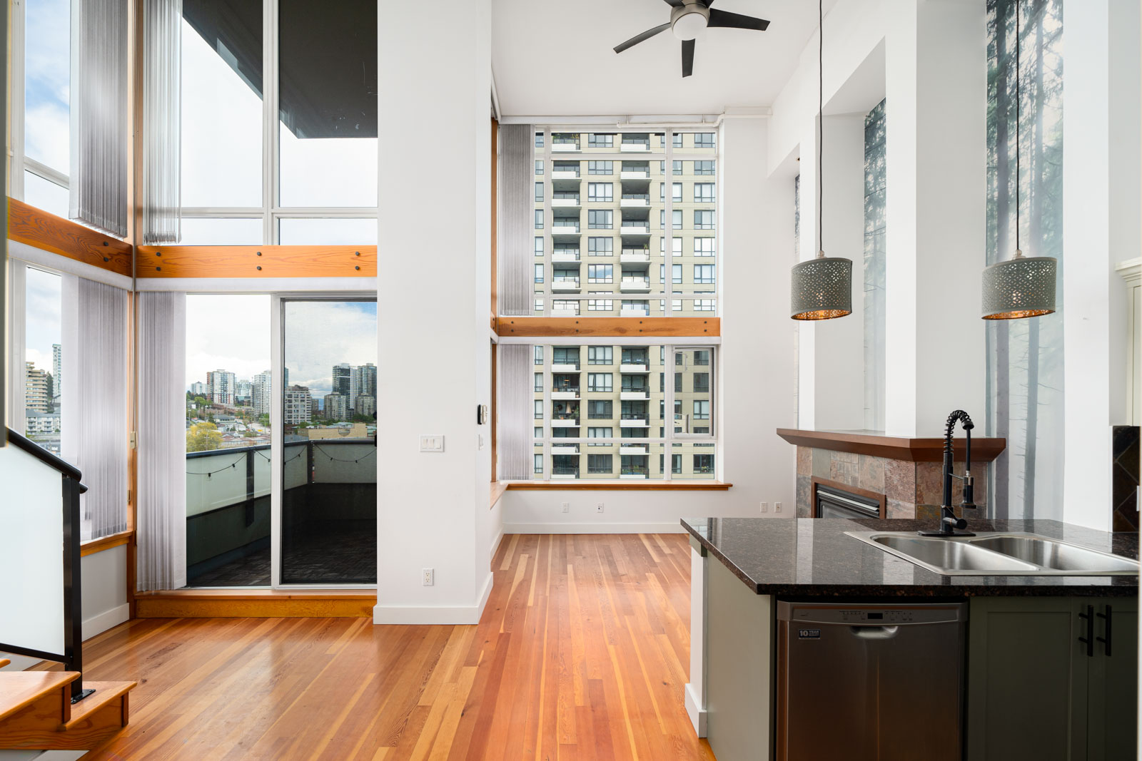Modern apartment interior with high ceilings, large windows, hardwood floors, a kitchen island with sink, and balcony overlooking city buildings.