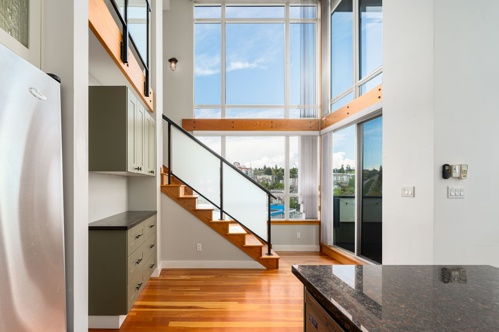 Modern loft-style interior with large windows, wooden floors, a staircase with frosted glass railing, and natural light filling the space.